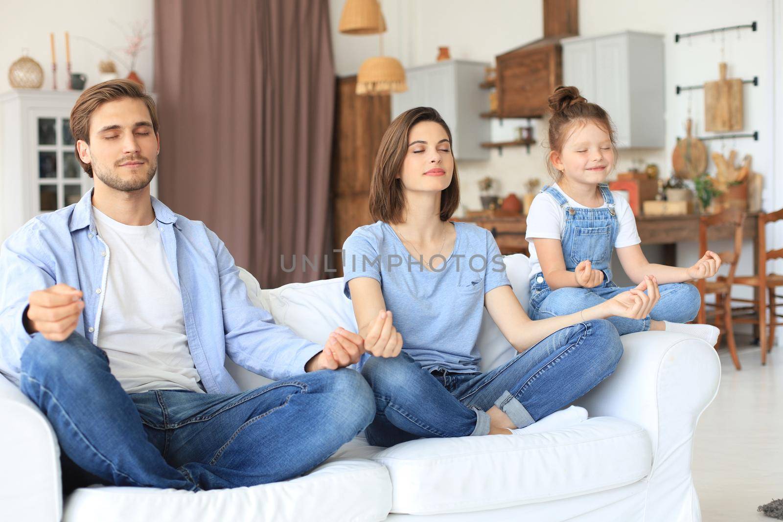 Calm young family with little daughter sit on couch practice yoga together, happy parents with small preschooler girl child rest on sofa meditate relieve negative emotions on weekend at home