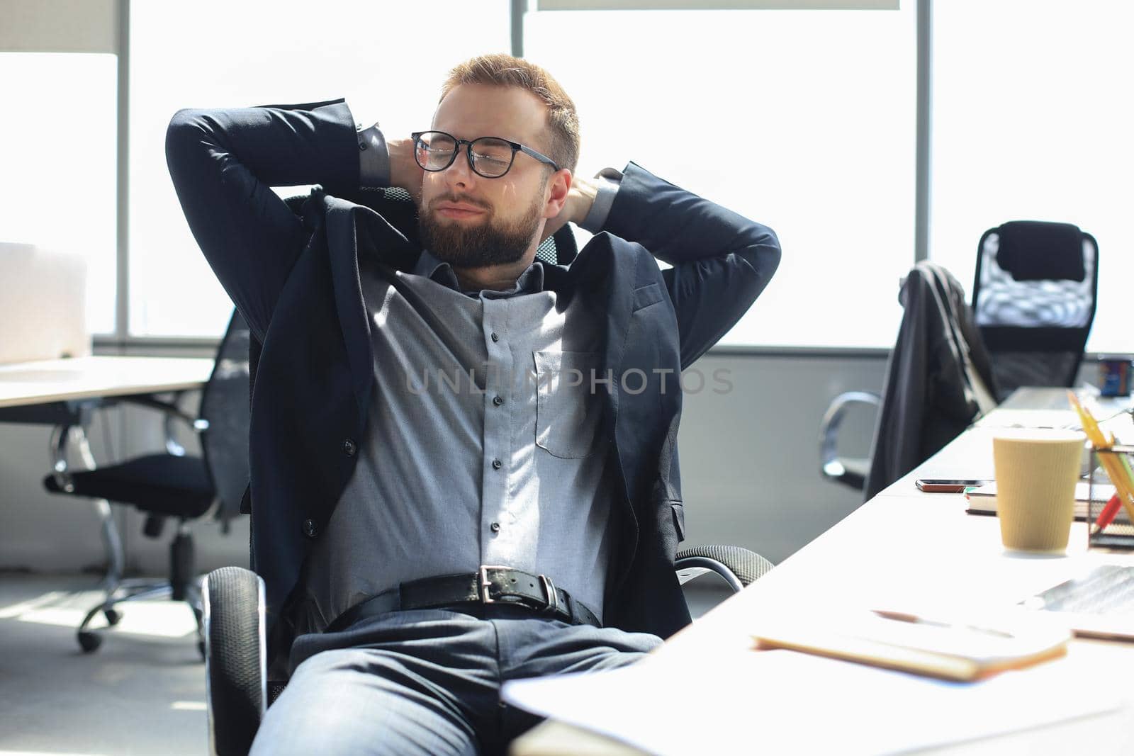 Young modern businessman keeping hands behind head and smiling while sitting in the office