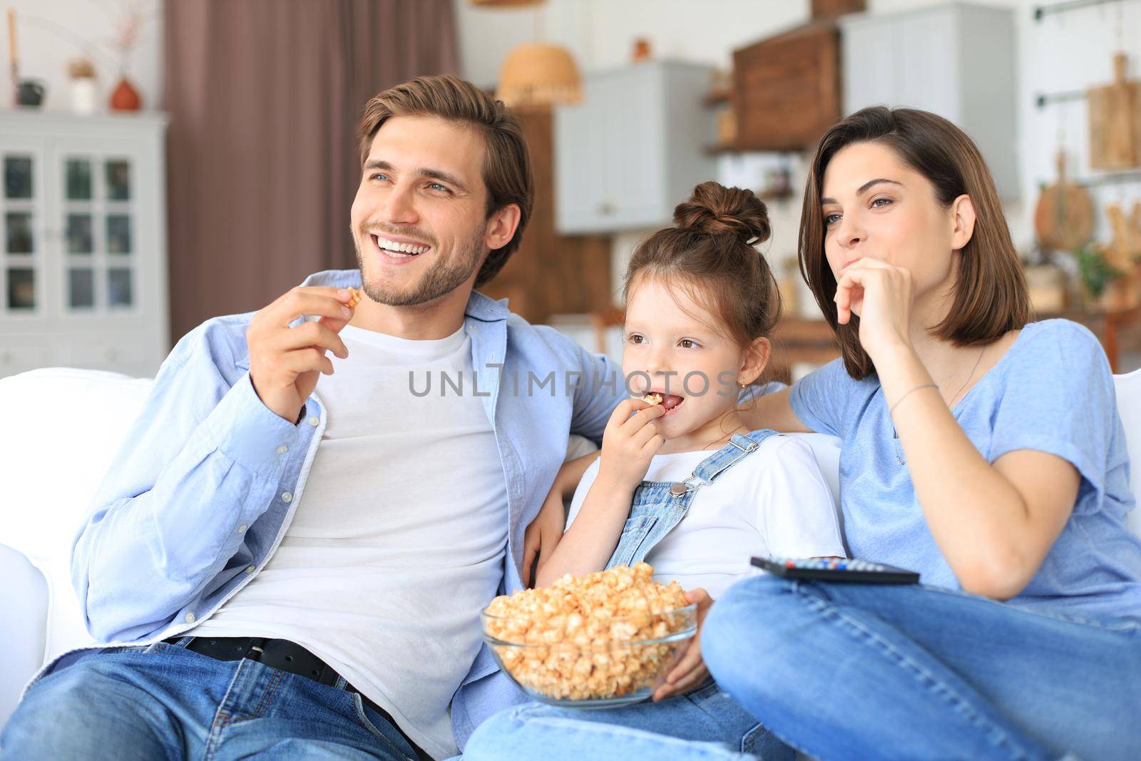 Happy family with child sitting on sofa watching tv and eating popcorn, young parents embracing daughter relaxing on couch together