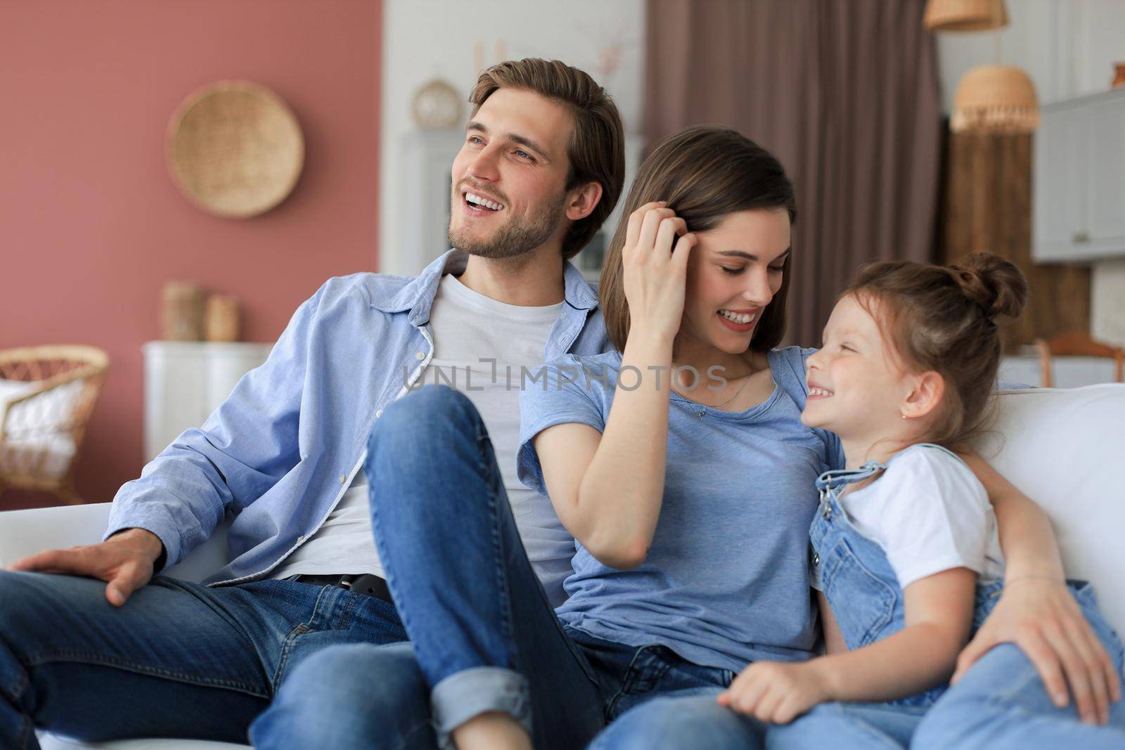 Positive friendly young parents with smiling little daughter sitting on sofa together while relaxing at home on weekend
