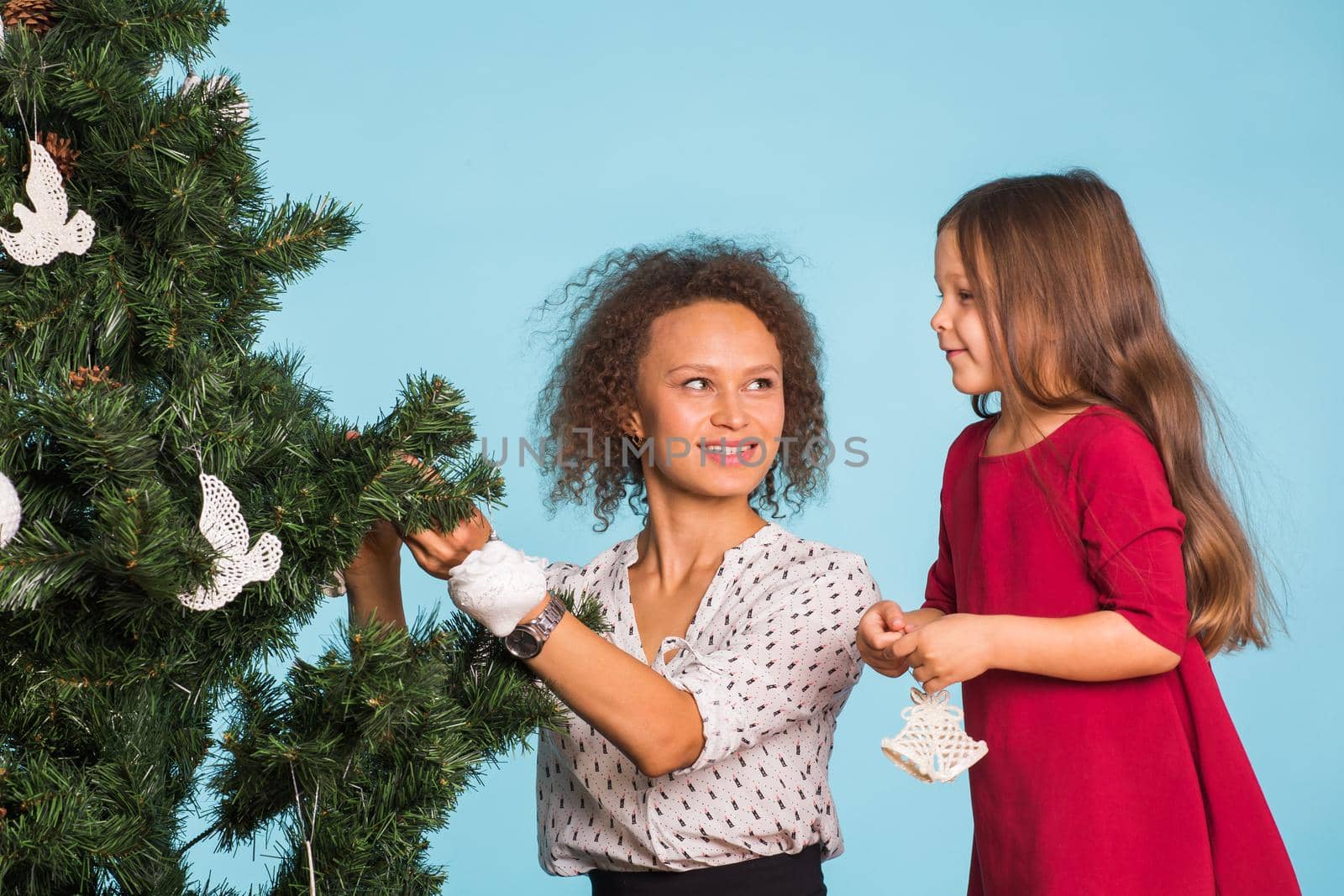 Holidays, family and christmas concept - pretty girl with mum are decorating a christmas tree on pink background.