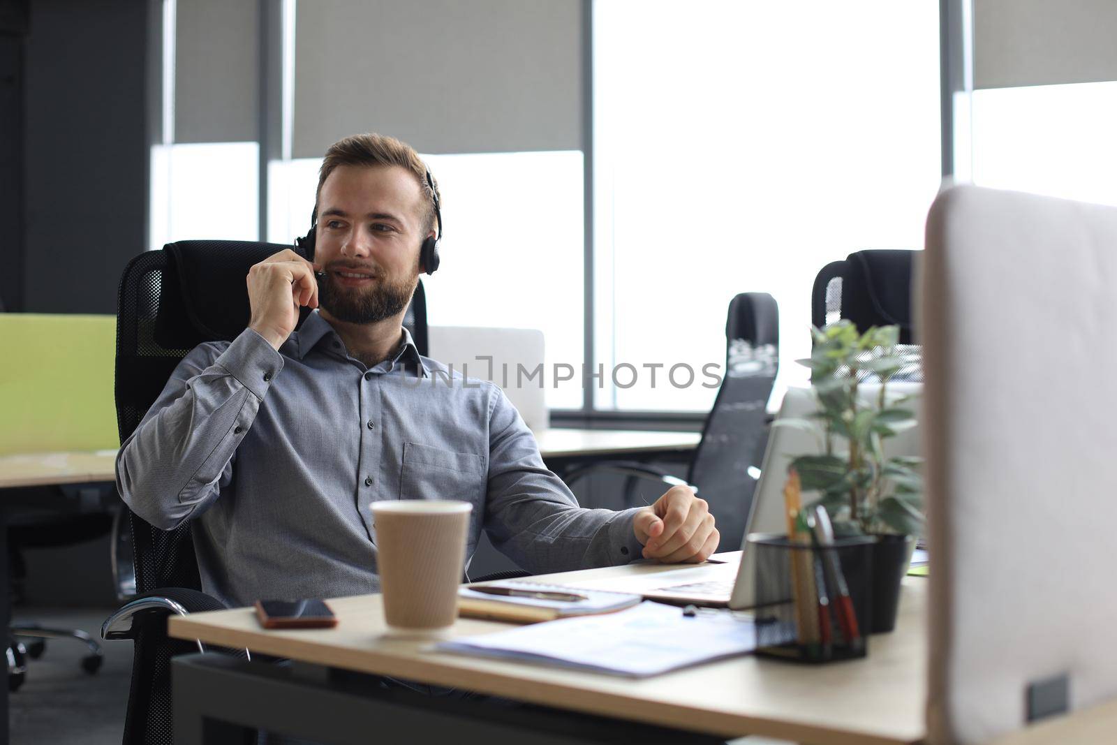 Smiling male call-center operator with headphones sitting at modern office, consulting online information in a laptop, looking up information in a file in order to be of assistance to the client