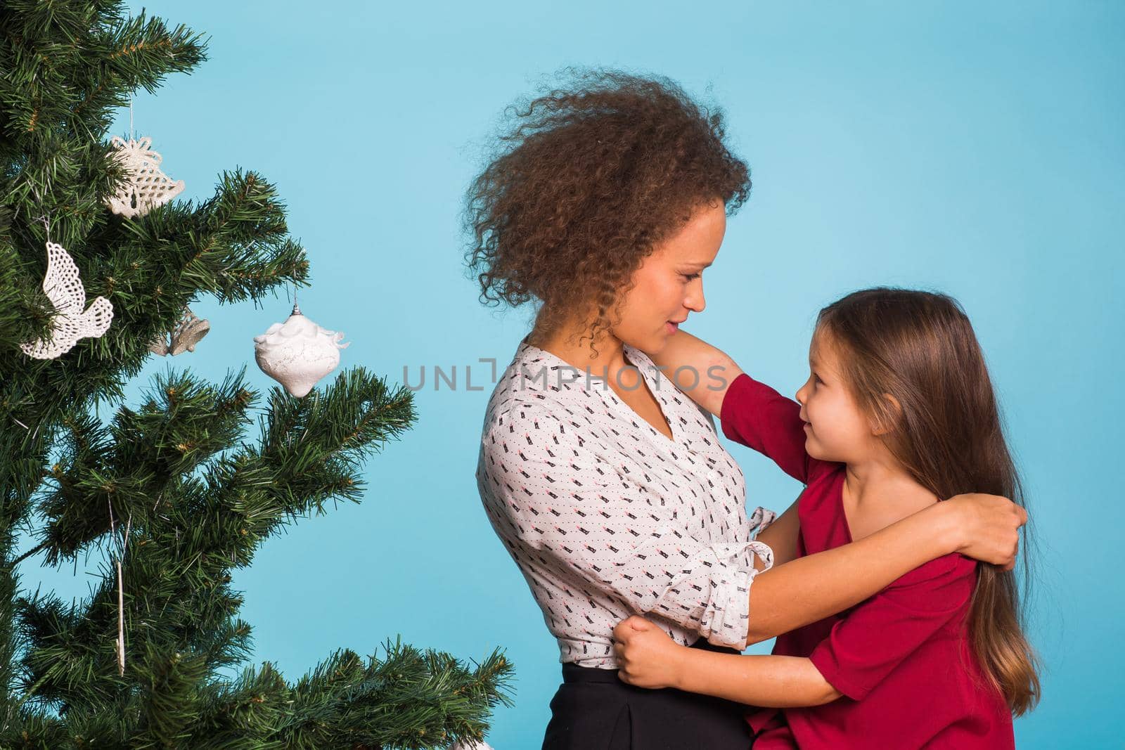 Mixed race mom and her daughter with Christmas tree on blue background by Satura86