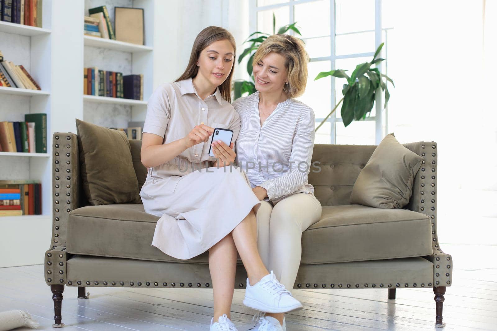 Middle aged mother and adult daughter hugging, using phone together, watching video or photos, sitting on cozy couch at home. by tsyhun