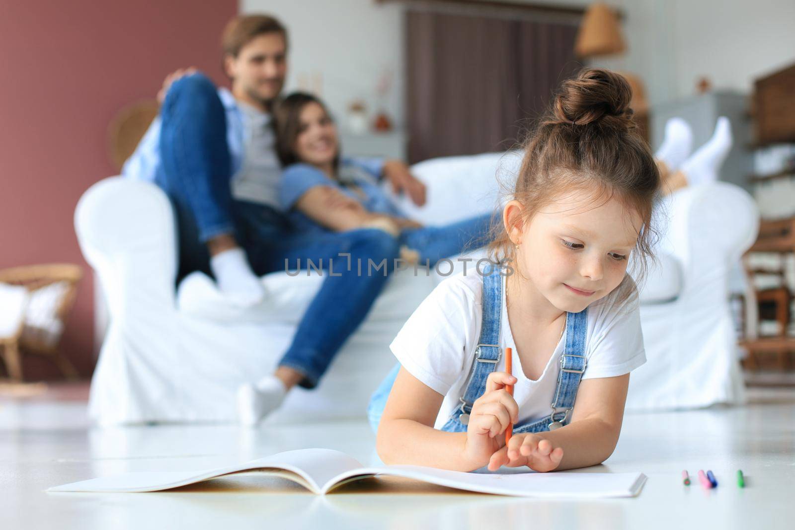 Smiling happy family sit relax on couch in living room watch little daughter drawing in album with colorful pencils. Happy weekends at home