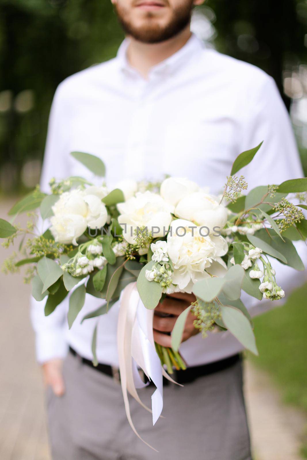 Caucasian groom wearing grey trousers and white shirt waiting for bride with bouquet of flowers. Concept of wedding photo session and floristic art.
