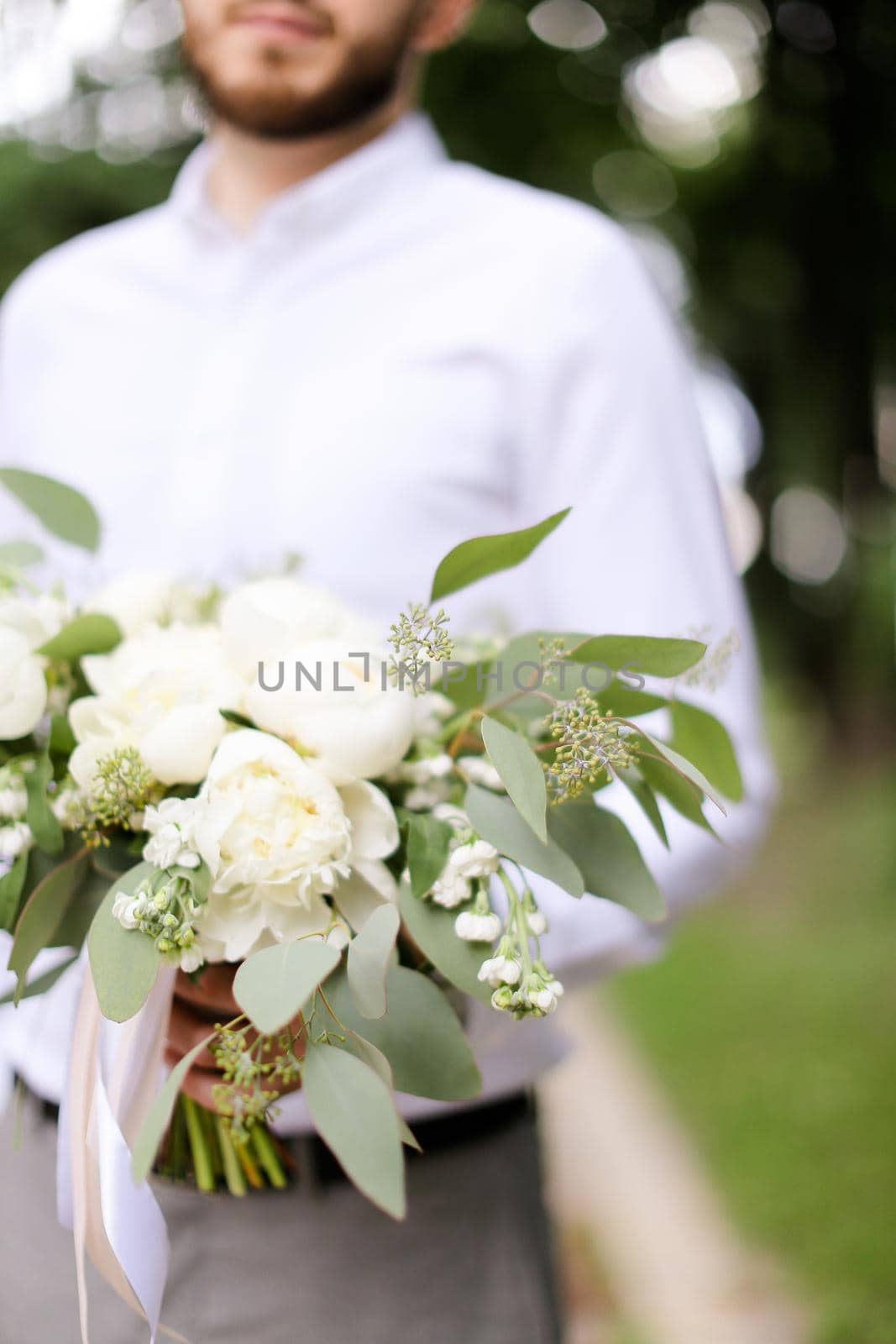 Caucasian groom wearing grey trousers and white shirt waiting for bride with bouquet, focus on flowers. Concept of wedding photo session and floristic art.