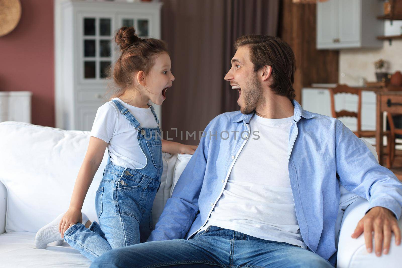 Smiling young father lying on couch at living room and play with happy little daughter