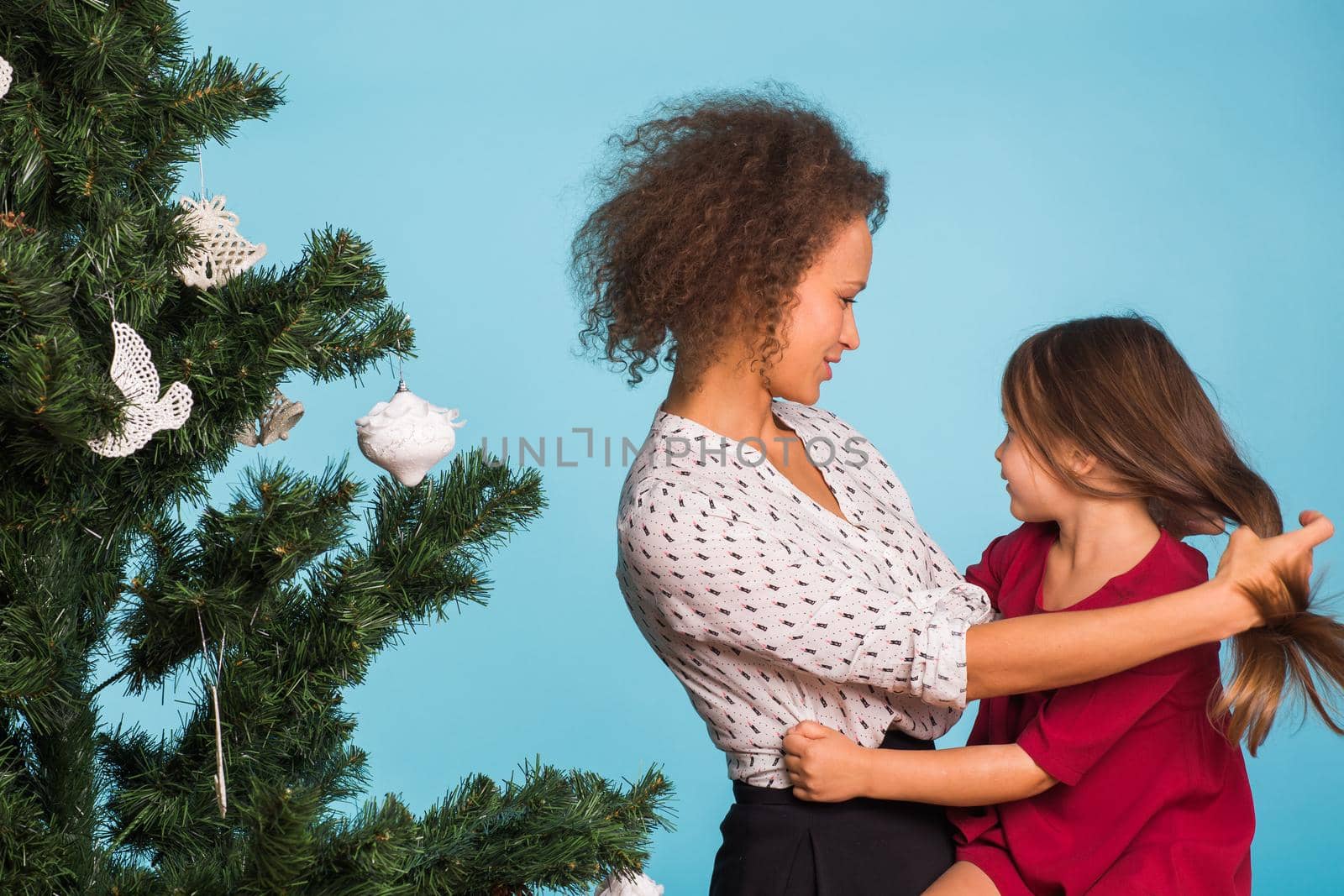 Mixed race mom and her daughter with Christmas tree on blue background by Satura86