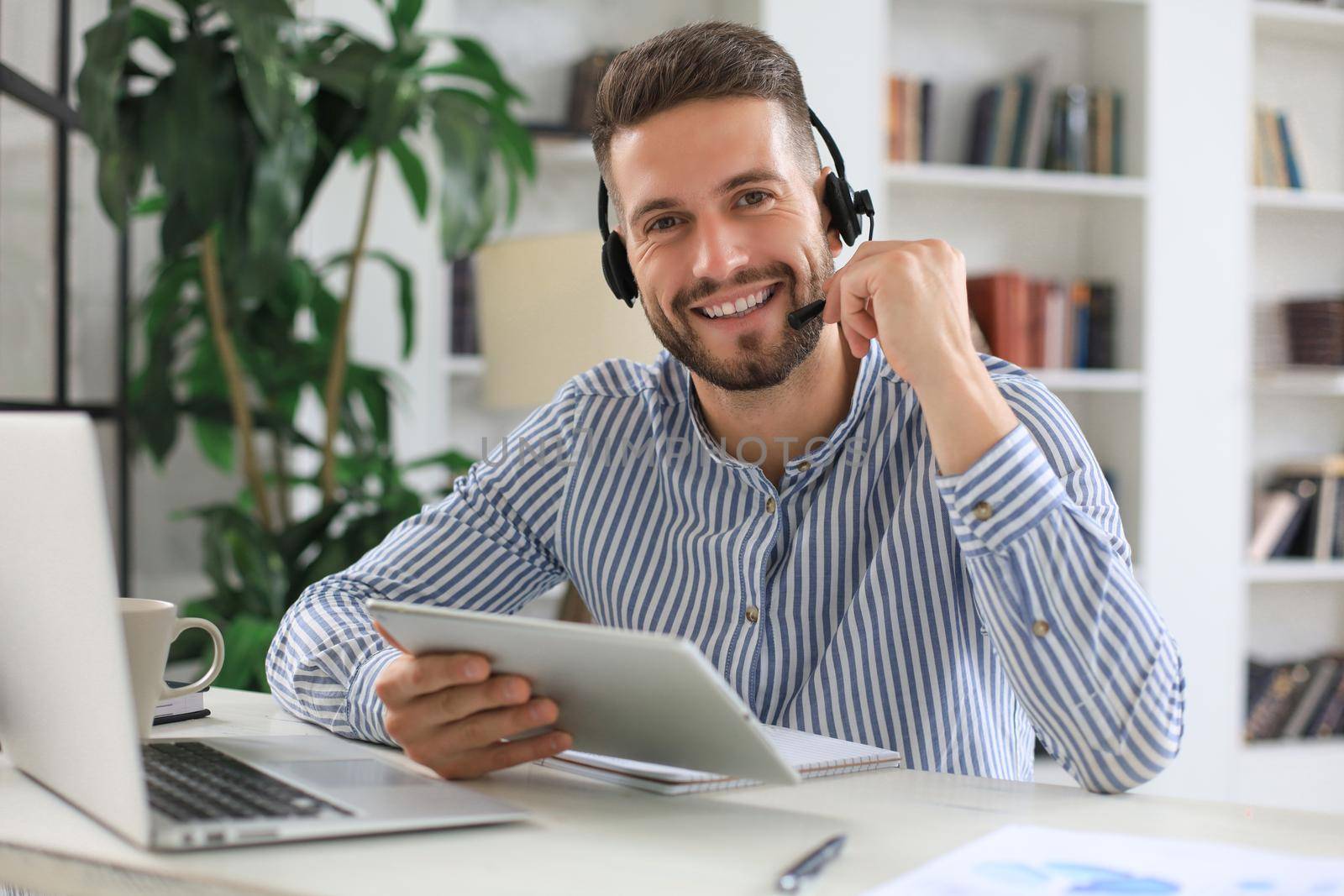 Cheerful young support phone male operator in headset, at workplace while using digital tablet, help service and client consulting call center concept.