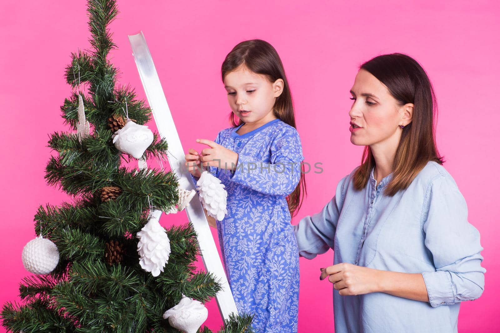 Holidays, family and christmas concept - pretty girl with mum are decorating a christmas tree on pink background by Satura86