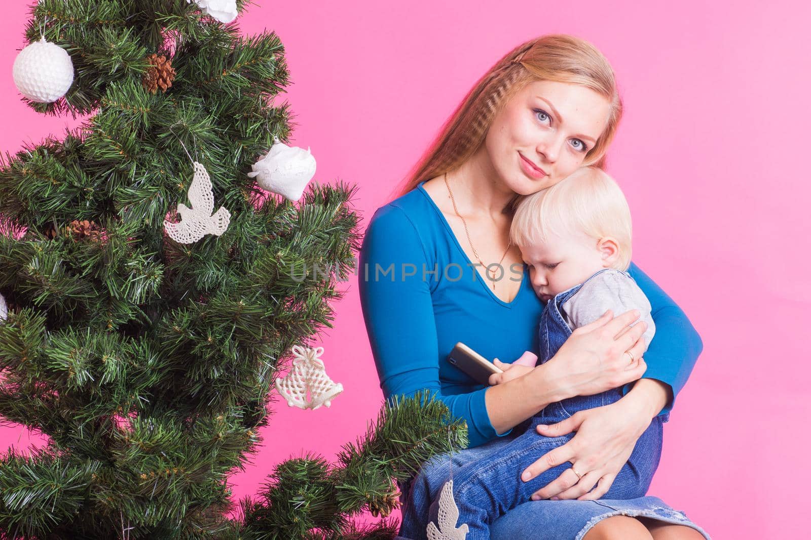 Christmas and holiday concept - Portrait of smiling woman holding her little daughter near Christmas tree on pink background.