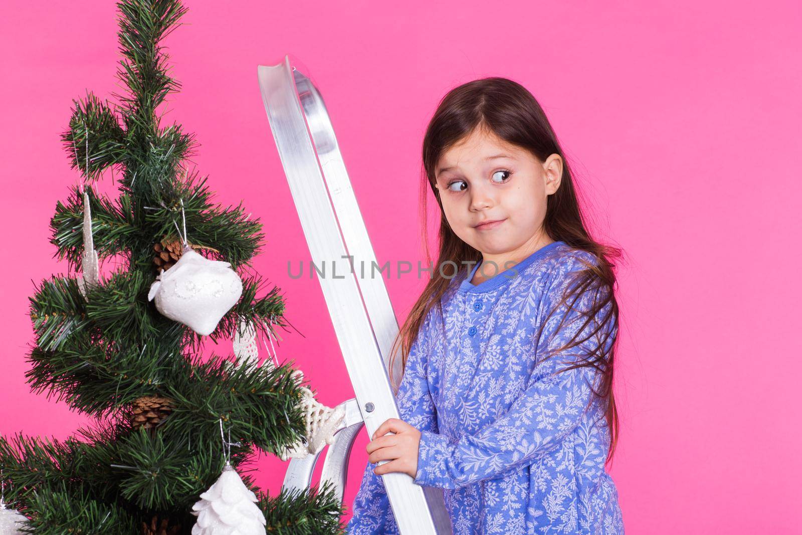 Children, holidays and christmas concept - little girl decorating christmas tree on pink background.