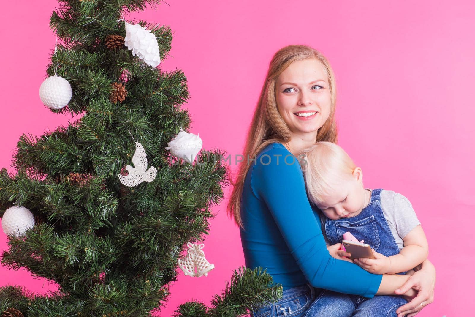 Christmas and holiday concept - Portrait of smiling woman holding her little daughter near Christmas tree on pink background.