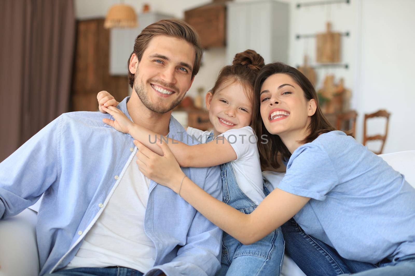 Positive friendly young parents with smiling little daughter sitting on sofa together while relaxing at home on weekend
