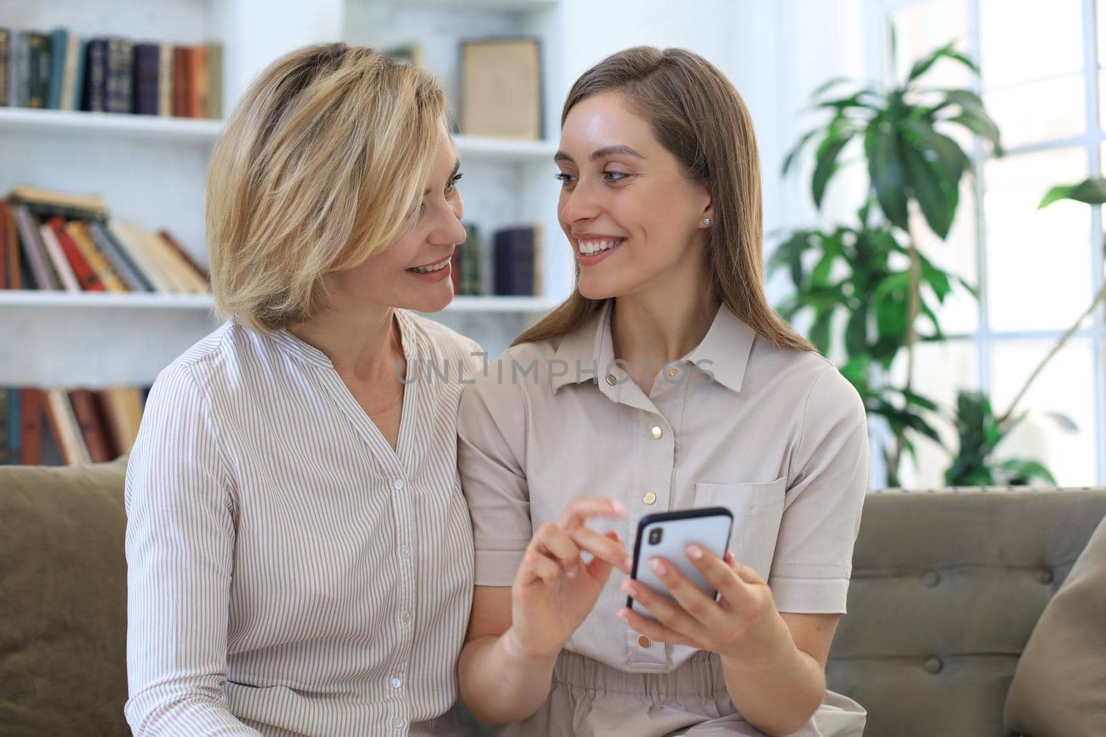 Middle aged mother and adult daughter hugging, using phone together, watching video or photos, sitting on cozy couch at home. by tsyhun