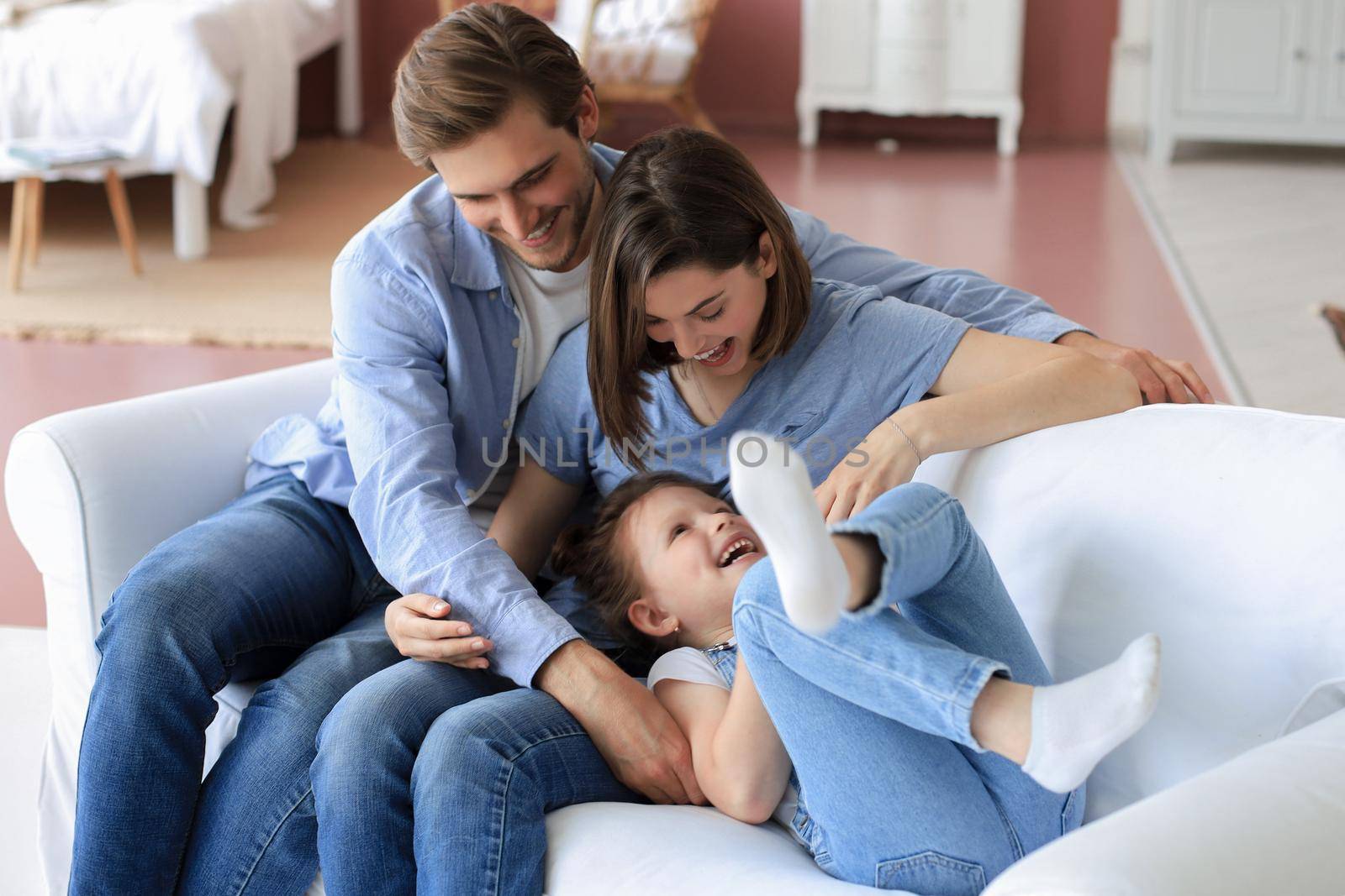 Positive friendly young parents with smiling little daughter sitting on sofa and having fun together at home