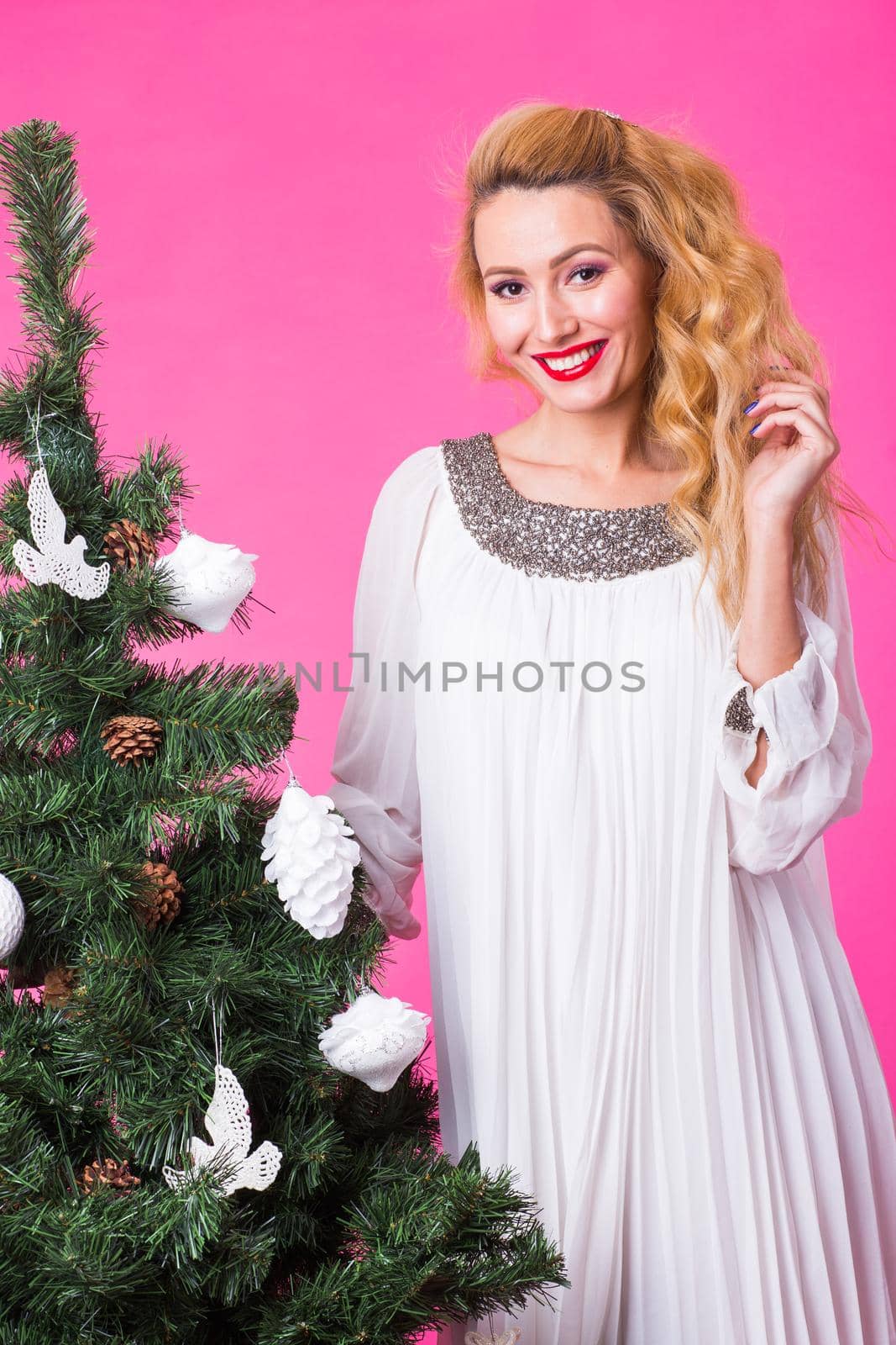 People, holidays and christmas concept - young blonde woman standing near christmas tree on pink background.