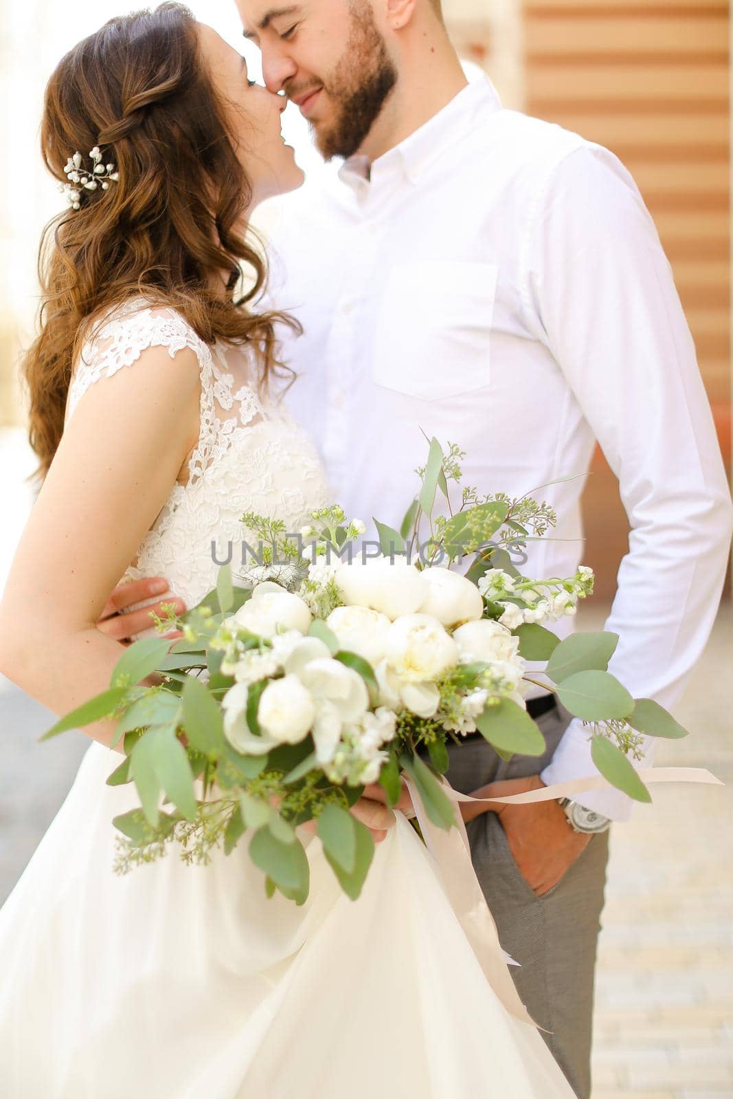 Happy american groom hugging bride keeping flowers and wearing white dress. Concept of married couple and love, wedding photo session.