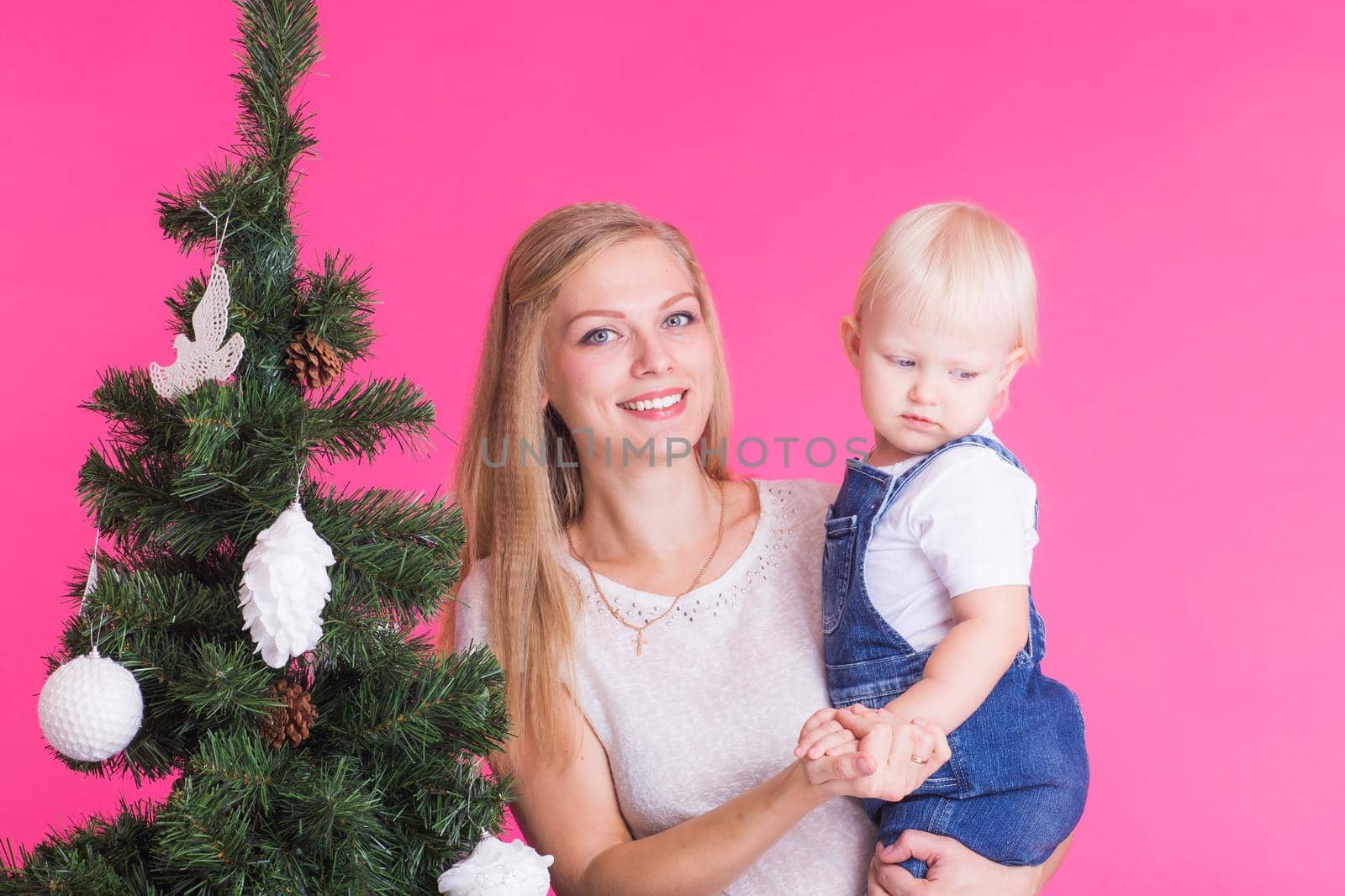 Christmas and holiday concept - Portrait of smiling woman holding her little daughter near Christmas tree on pink background.