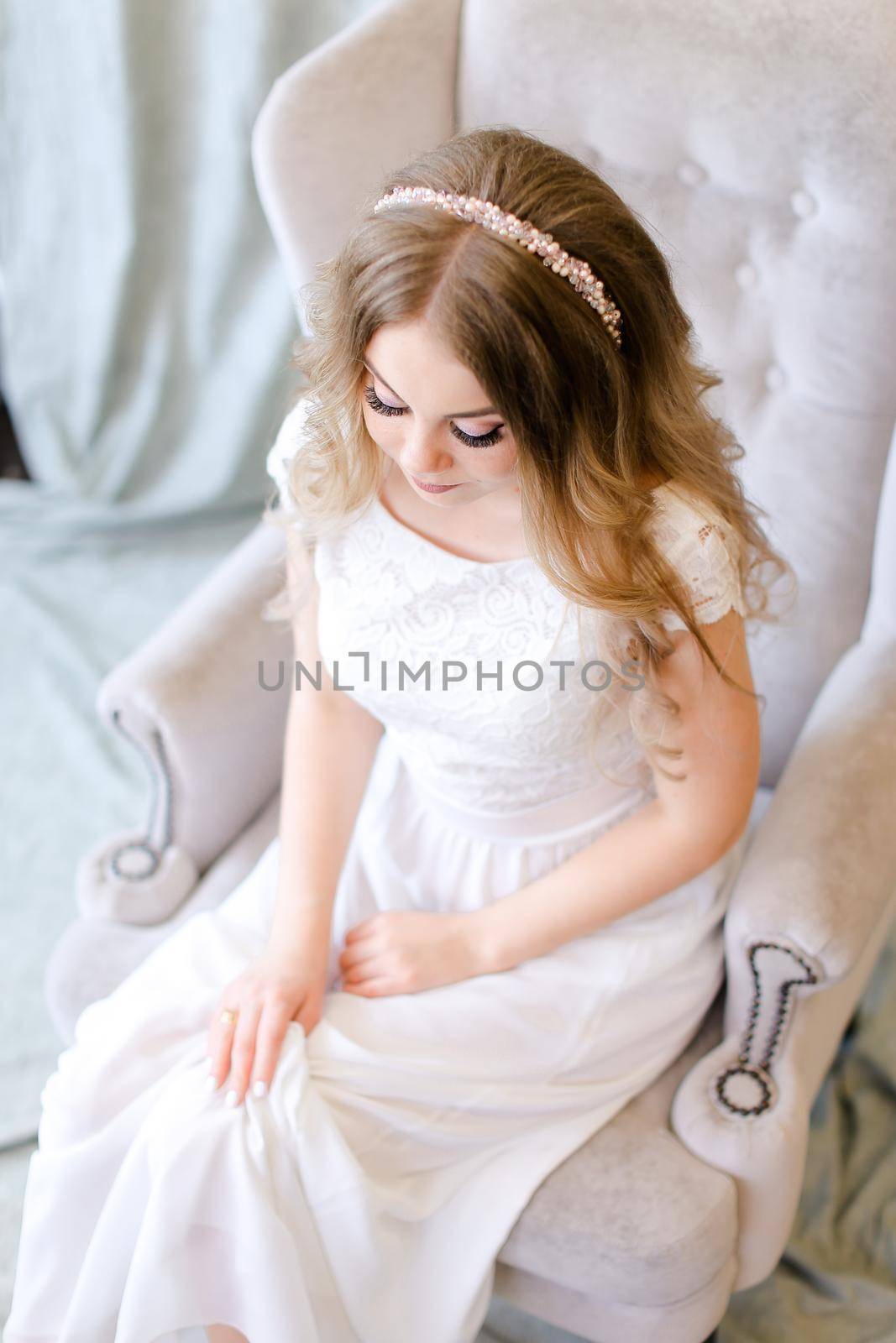 Nice fiancee sitting in arm chair at studio. Concept of bridal photo session and wedding fashion.