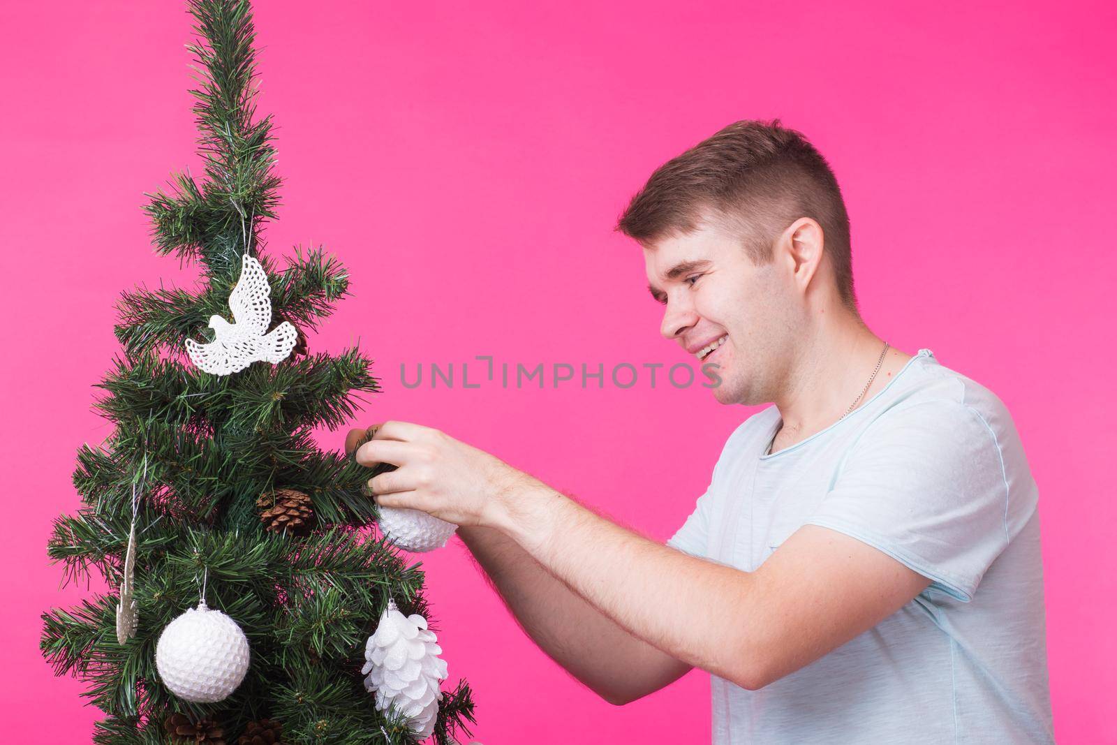 People, holidays and christmas concept - young man decorating christmas tree on pink background.
