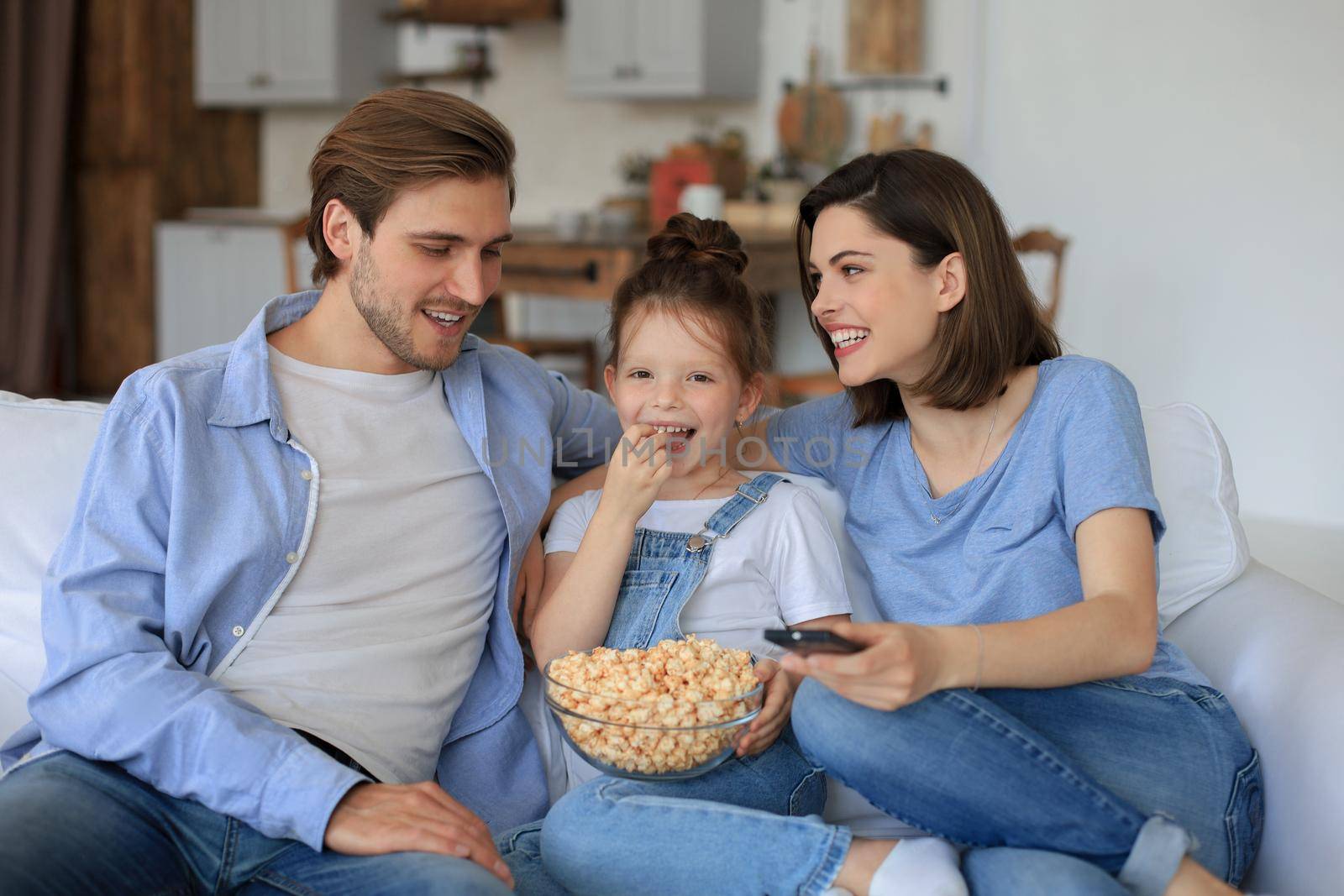 Happy family with child sitting on sofa watching tv and eating popcorn, young parents embracing daughter relaxing on couch together