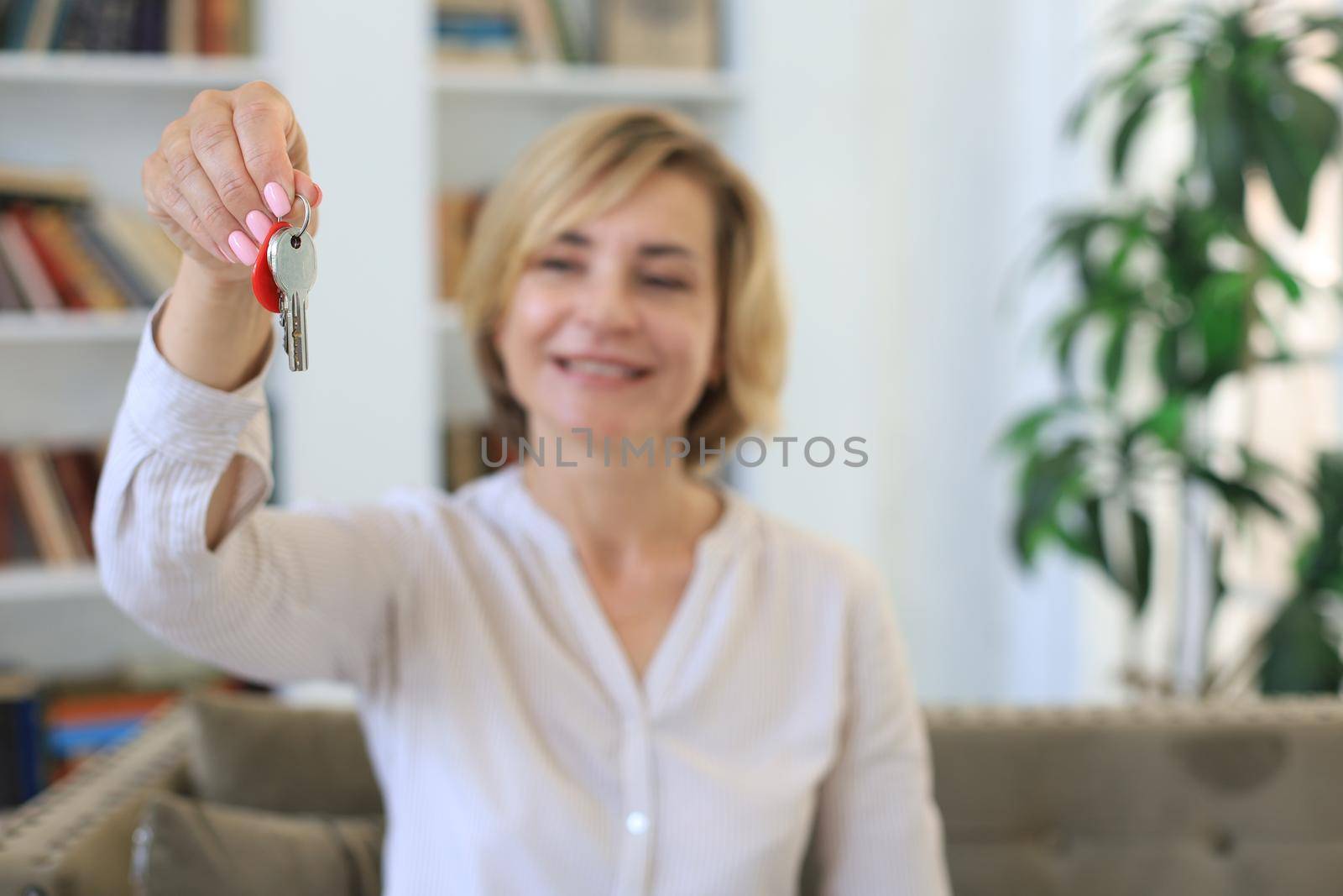 Cheerful middle aged female, real estate agent, sitting on couch, holding keys to new house