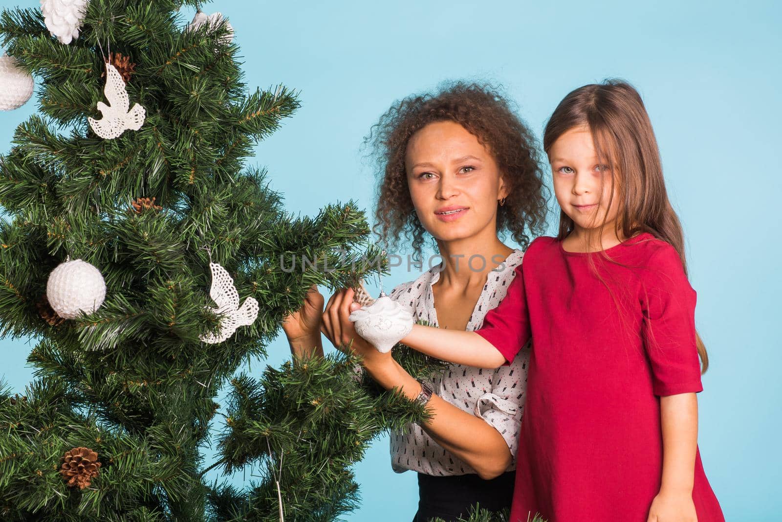 Holidays, family and christmas concept - mixed race mother and daughter decorating christmas tree on blue background by Satura86