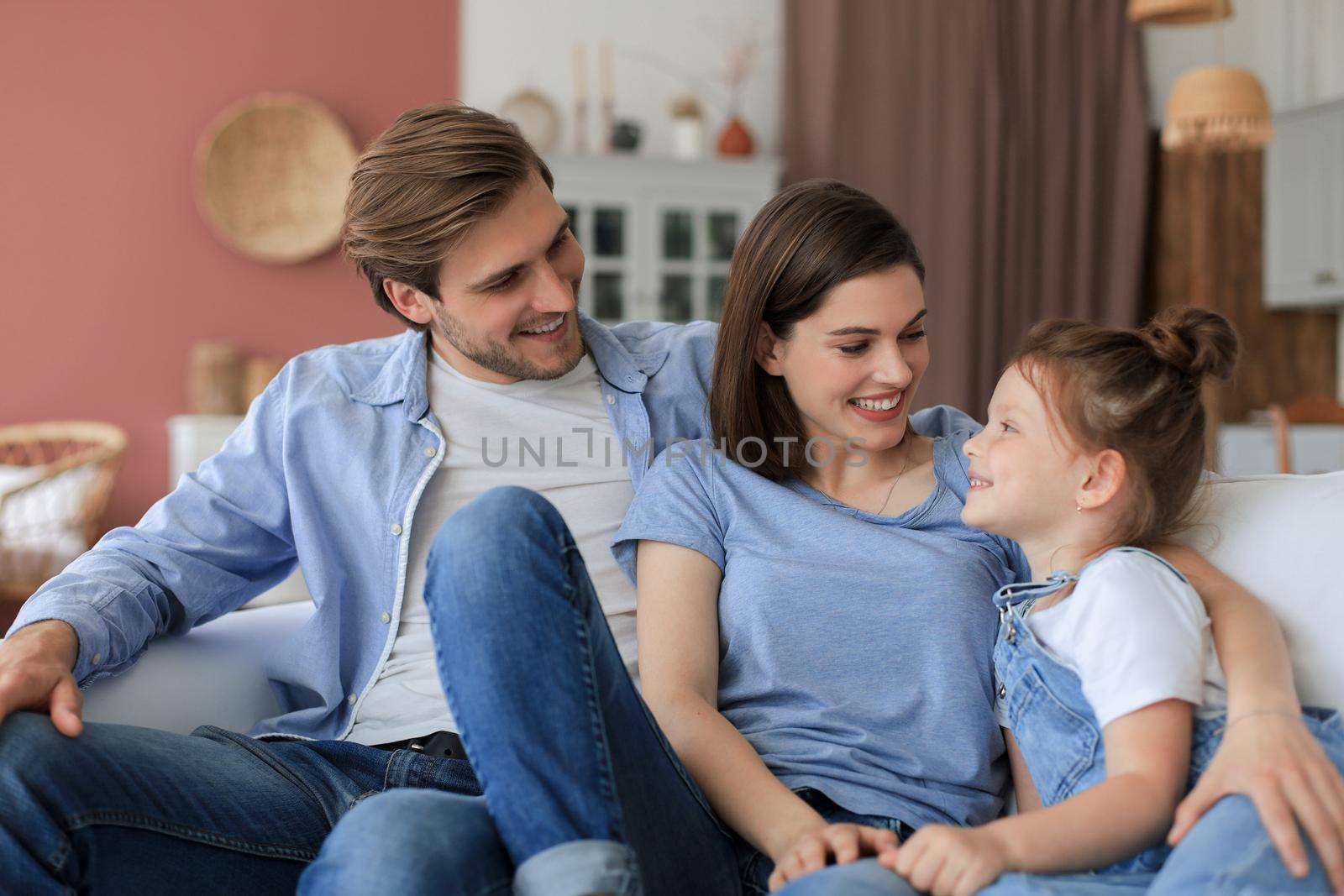 Positive friendly young parents with smiling little daughter sitting on sofa together while relaxing at home on weekend