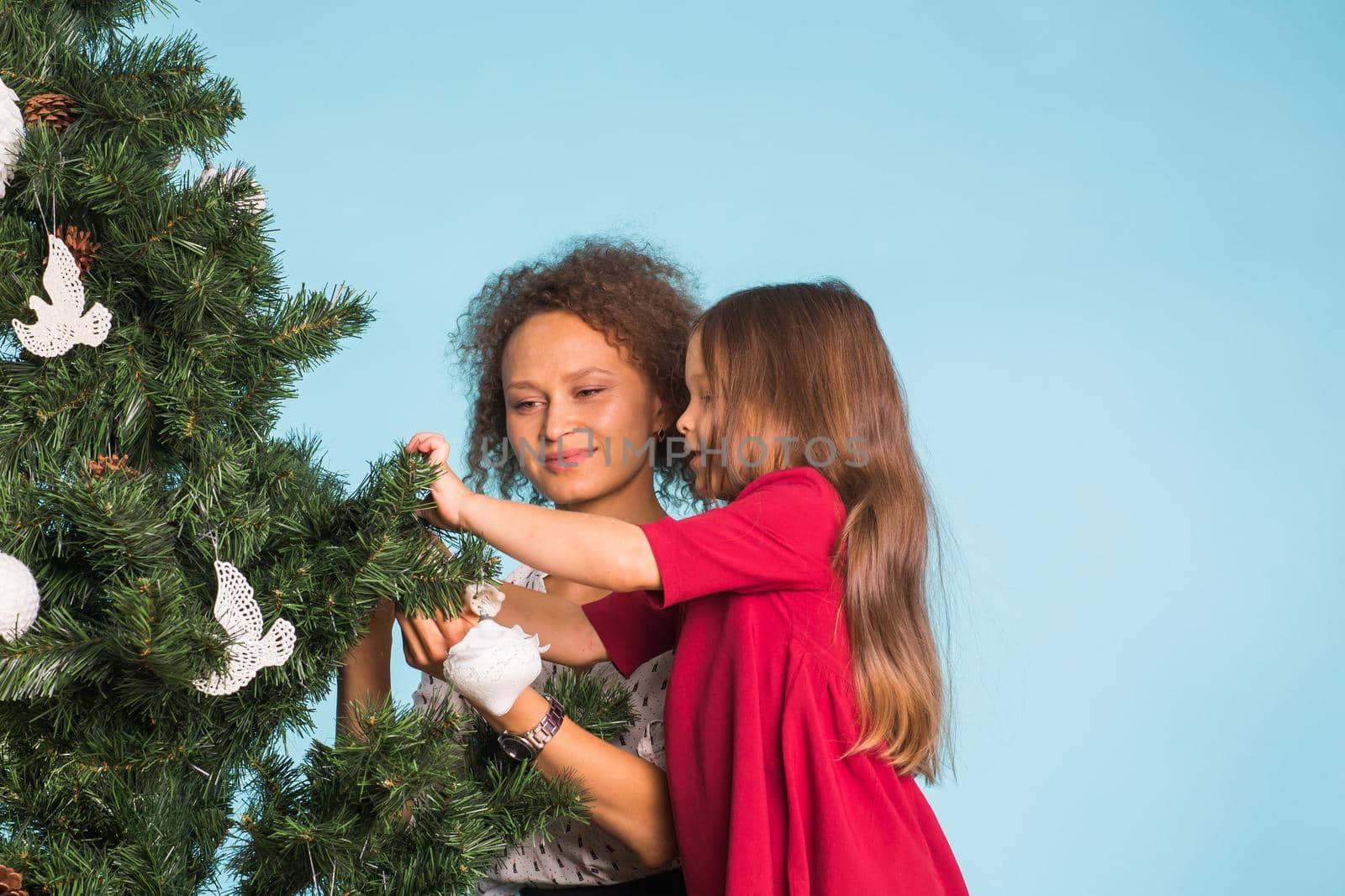 Holidays, family and christmas concept - pretty girl with mum are decorating a christmas tree on pink background.