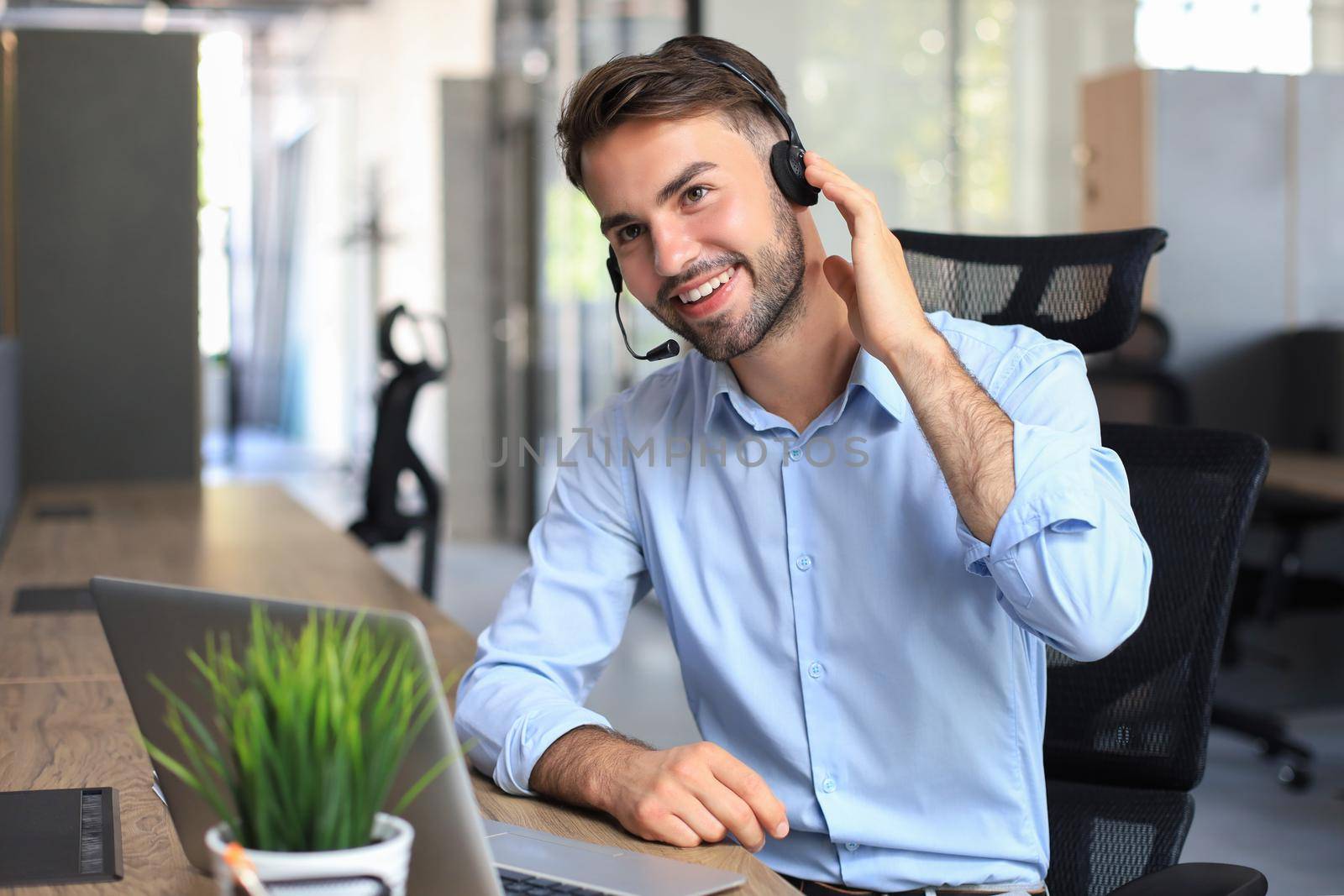 Smiling male call-center operator with headphones sitting at modern office, consulting online information in a laptop, looking up information in a file in order to be of assistance to the client