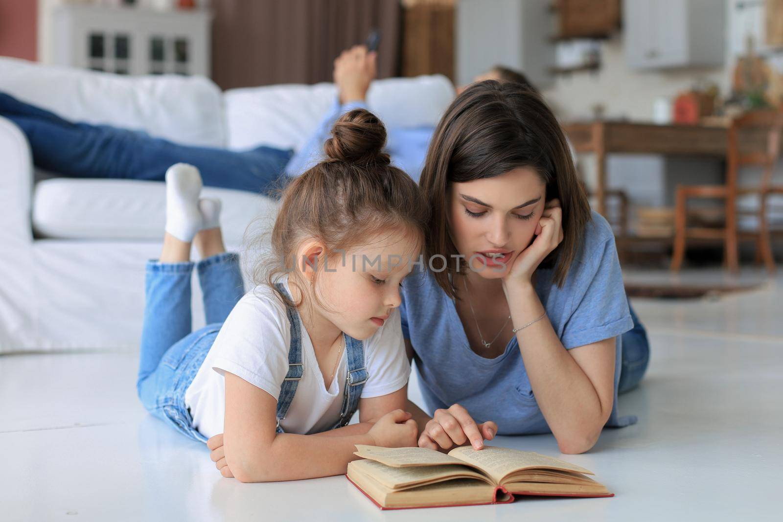 Mother and little daughter read a book together, lying on the floor in the living room