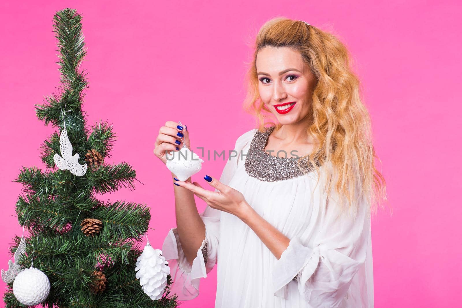 People, holidays and christmas concept - young blonde woman decorating christmas tree on pink background.