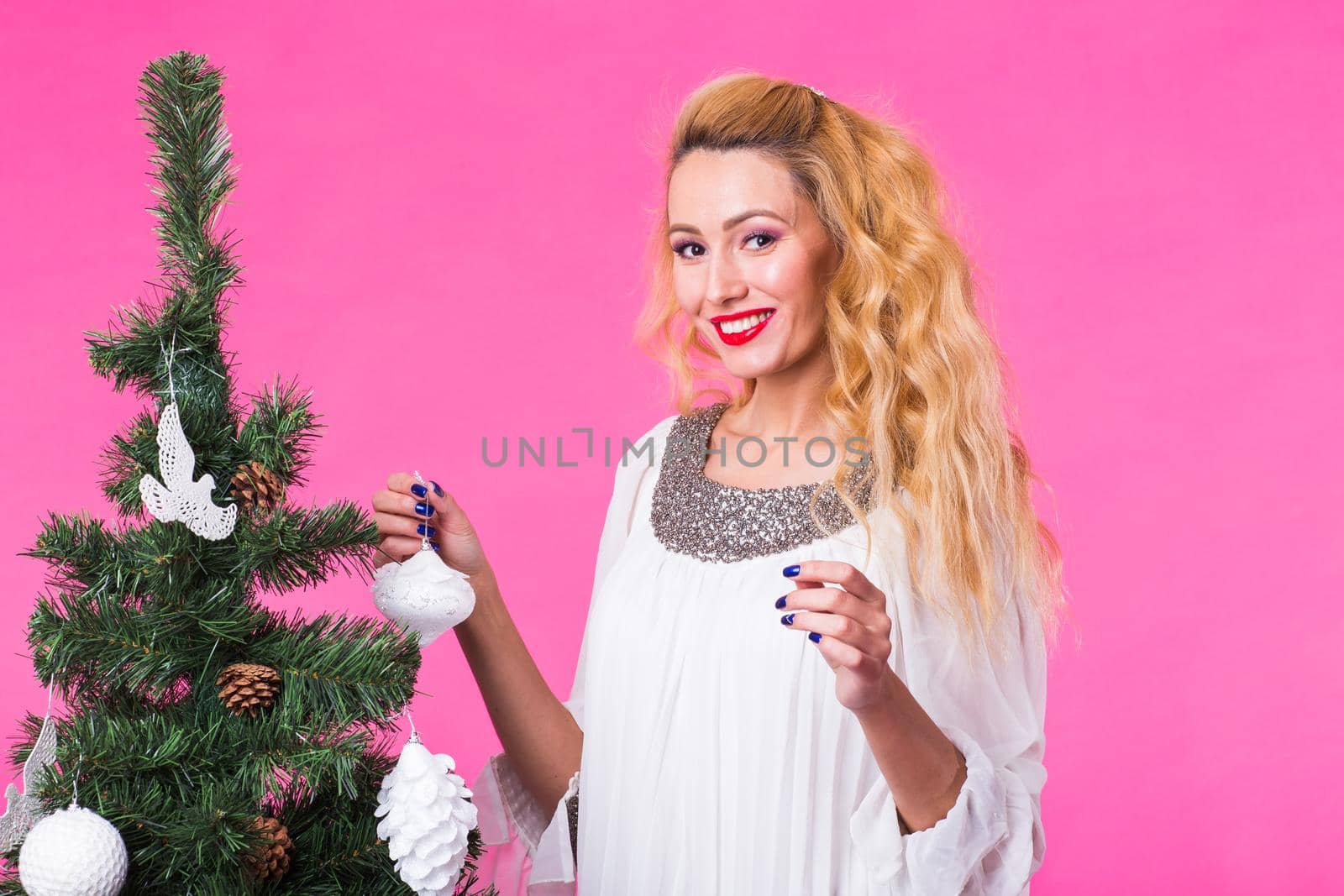 Christmas, holidays and people concept - young happy blonde woman decorating christmas tree on pink background by Satura86