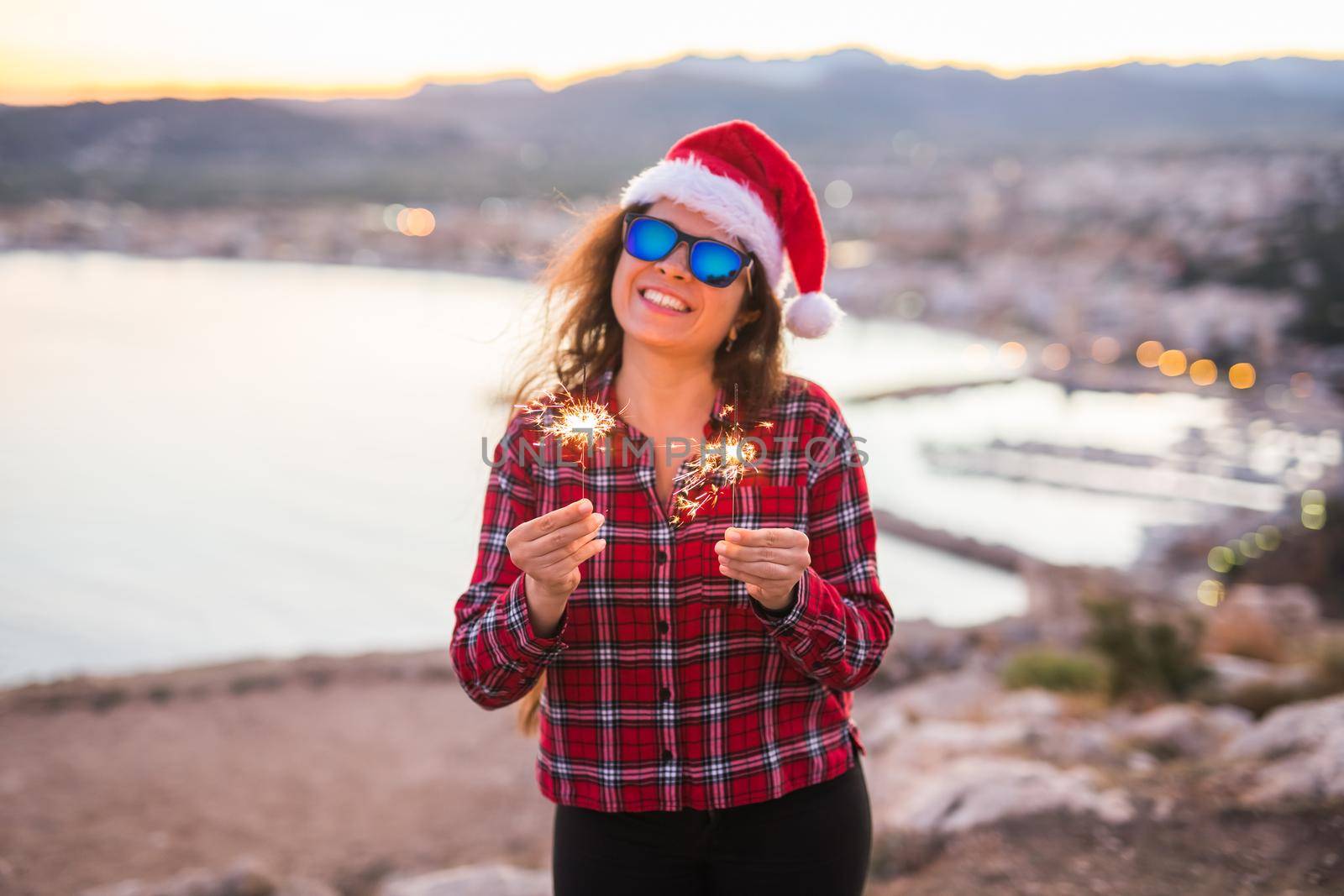 Christmas, holidays, people concept - woman in Santa hat and sunglasses holding sparklers over sea background by Satura86