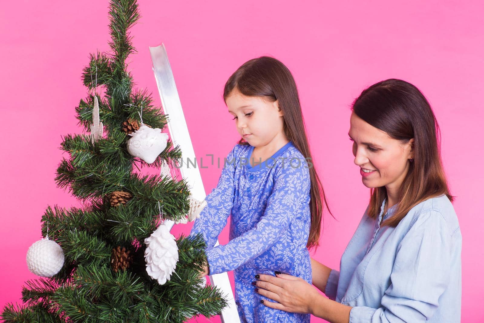 people, family and christmas concept - mother and daughter decorating christmas tree on pink background.