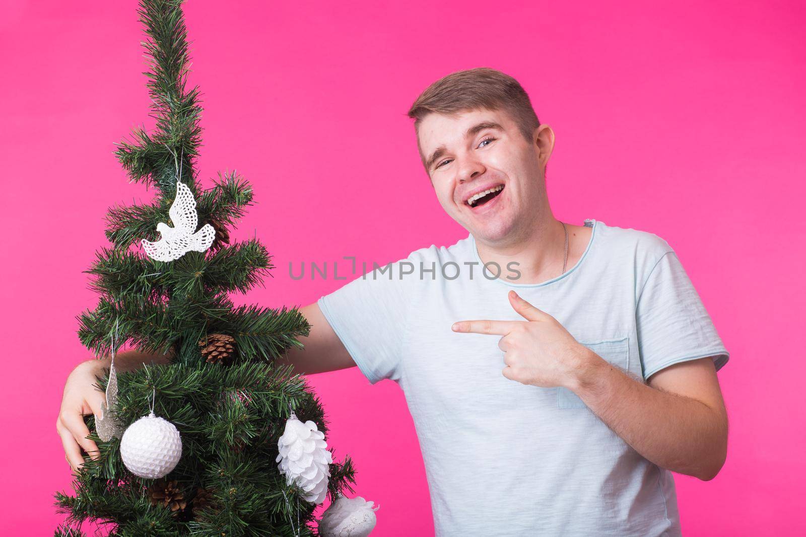 People, holidays and christmas concept - young man decorating christmas tree on pink background.