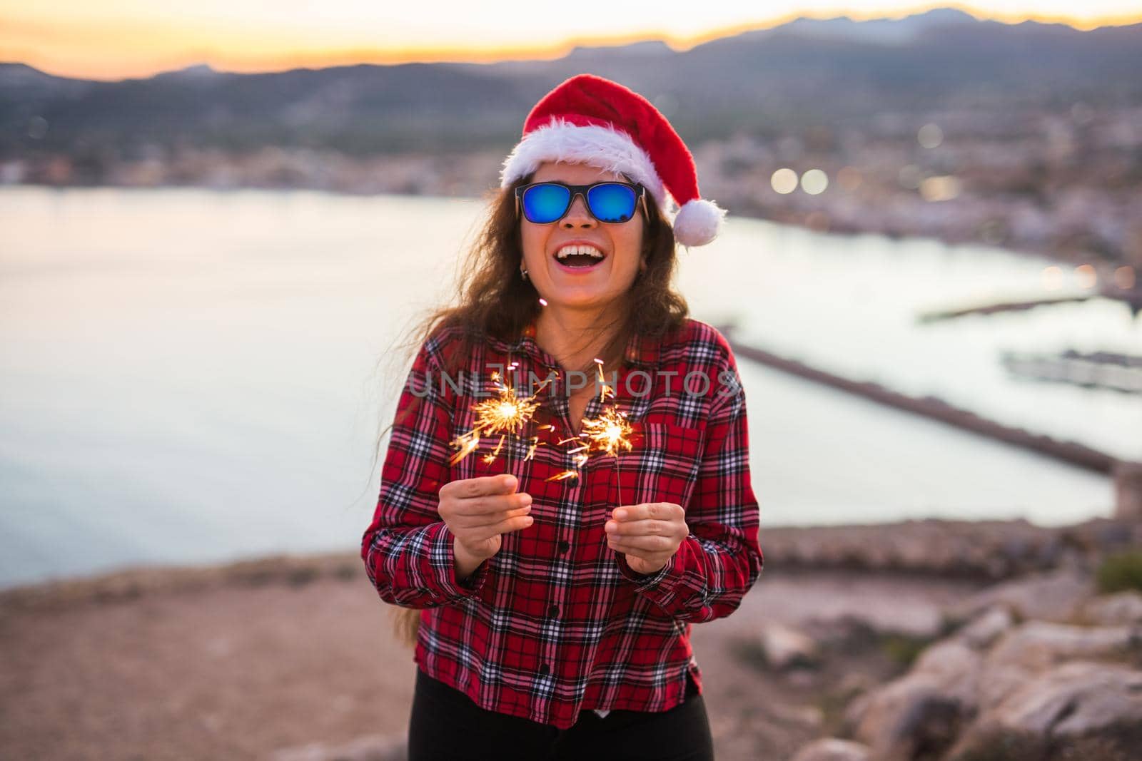 Holiday, Christmas and people concept - Young happy woman in Santa hat over beautiful landscape with fireworks and sparklers.