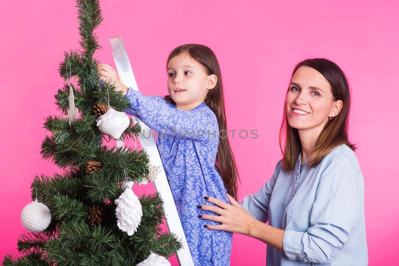 people, family and christmas concept - mother and daughter decorating christmas tree on pink background.