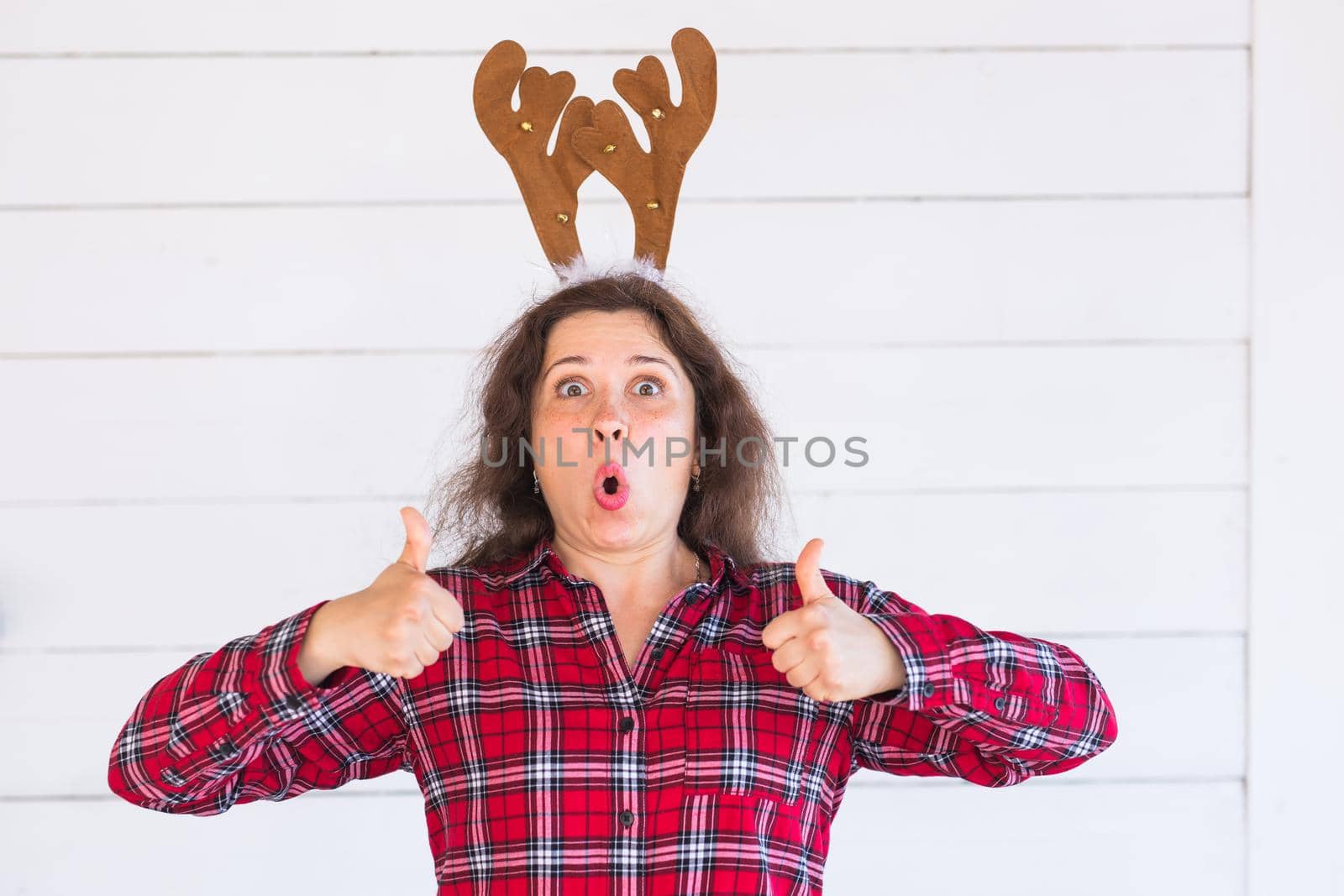 Happy beautiful woman in Christmas costume with deer horns on her head gesturing thumbs up over white background.