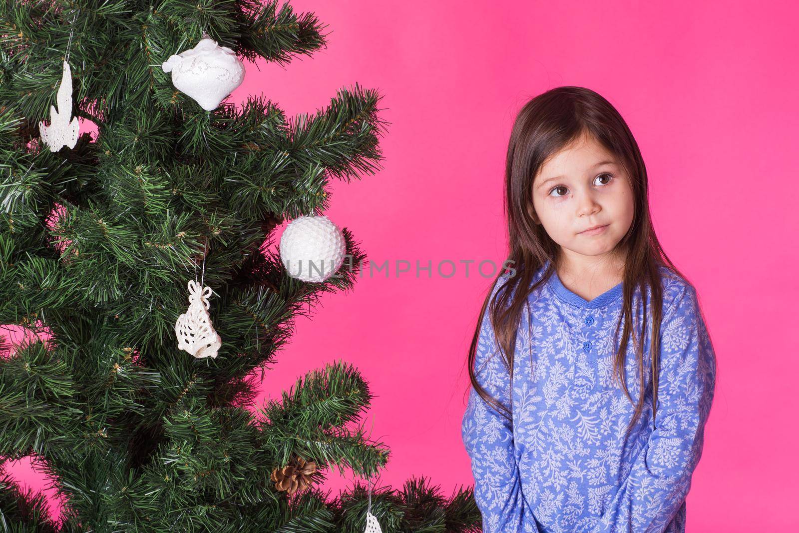 Children, holidays and christmas concept - little girl decorating christmas tree on pink background.