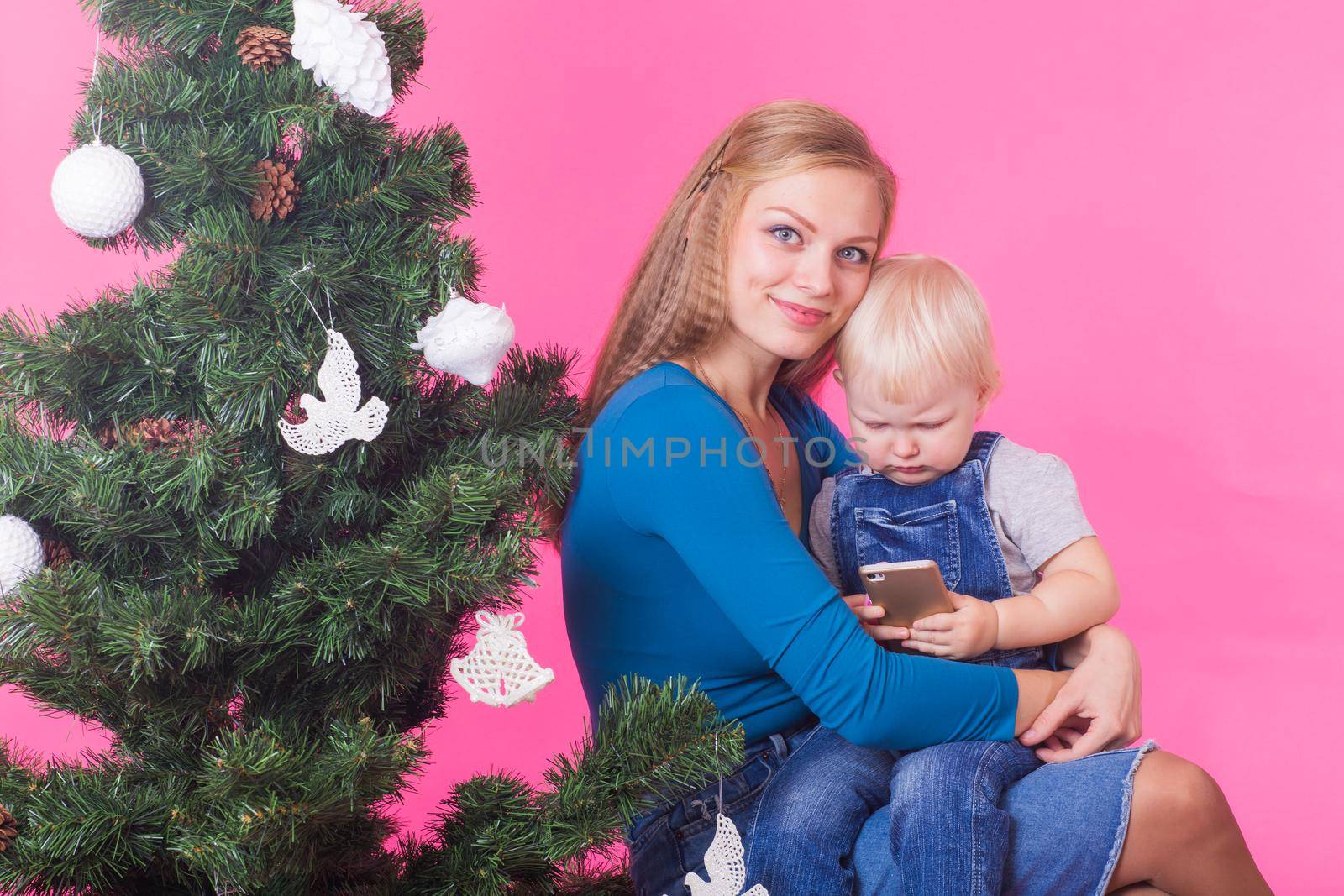 pHolidays, family and christmas concept - young woman with her baby near christmas tree on pink background.