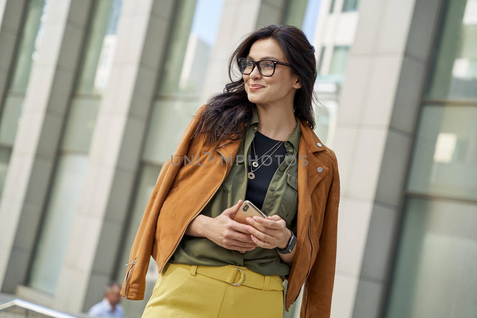 Young stylish woman holding smartphone, looking aside and smiling while standing against blurred urban background, enjoying walking on the city street by friendsstock