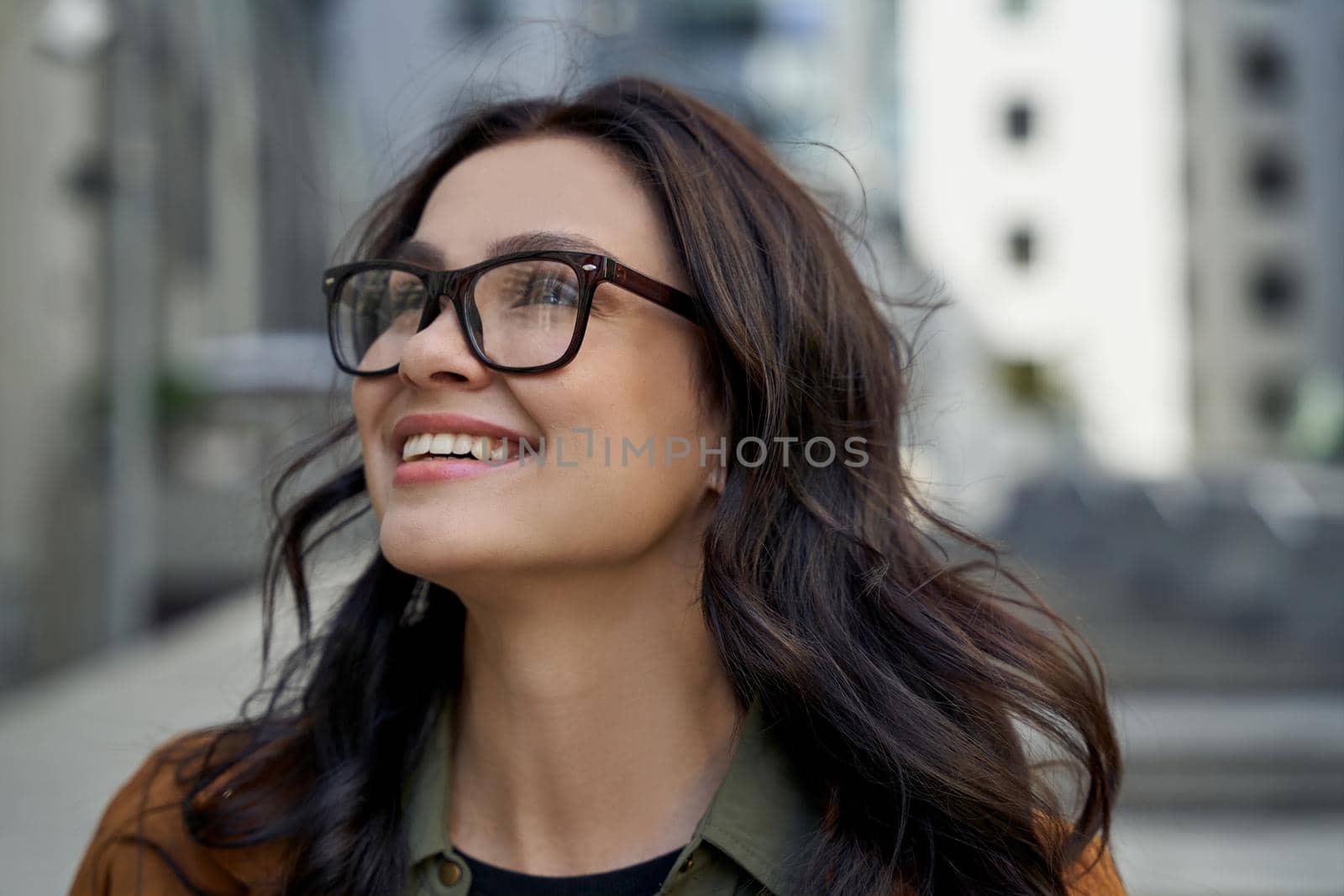 Close up portrait of a young happy charming woman wearing eyeglasses looking aside and smiling while walking city streets, standing against blurred urban background by friendsstock