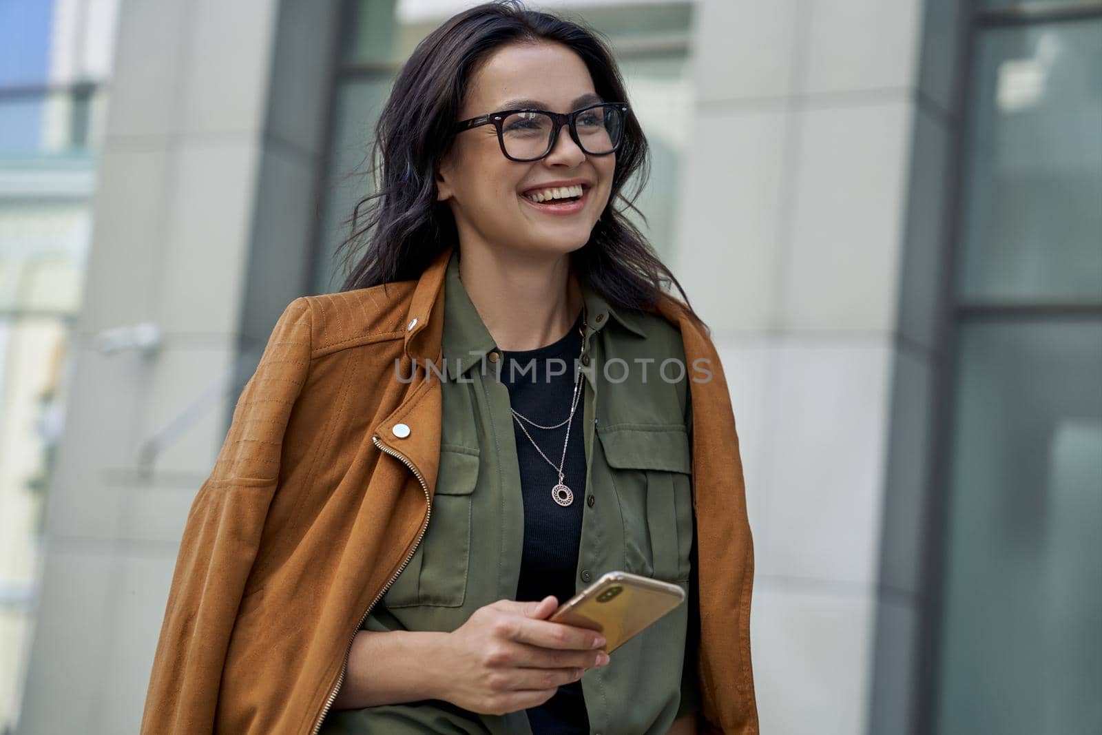 Being outside on a beautiful day. Young happy fashionable woman wearing eyeglasses walking on the city street, using her smartphone and smiling while standing against blurred urban background