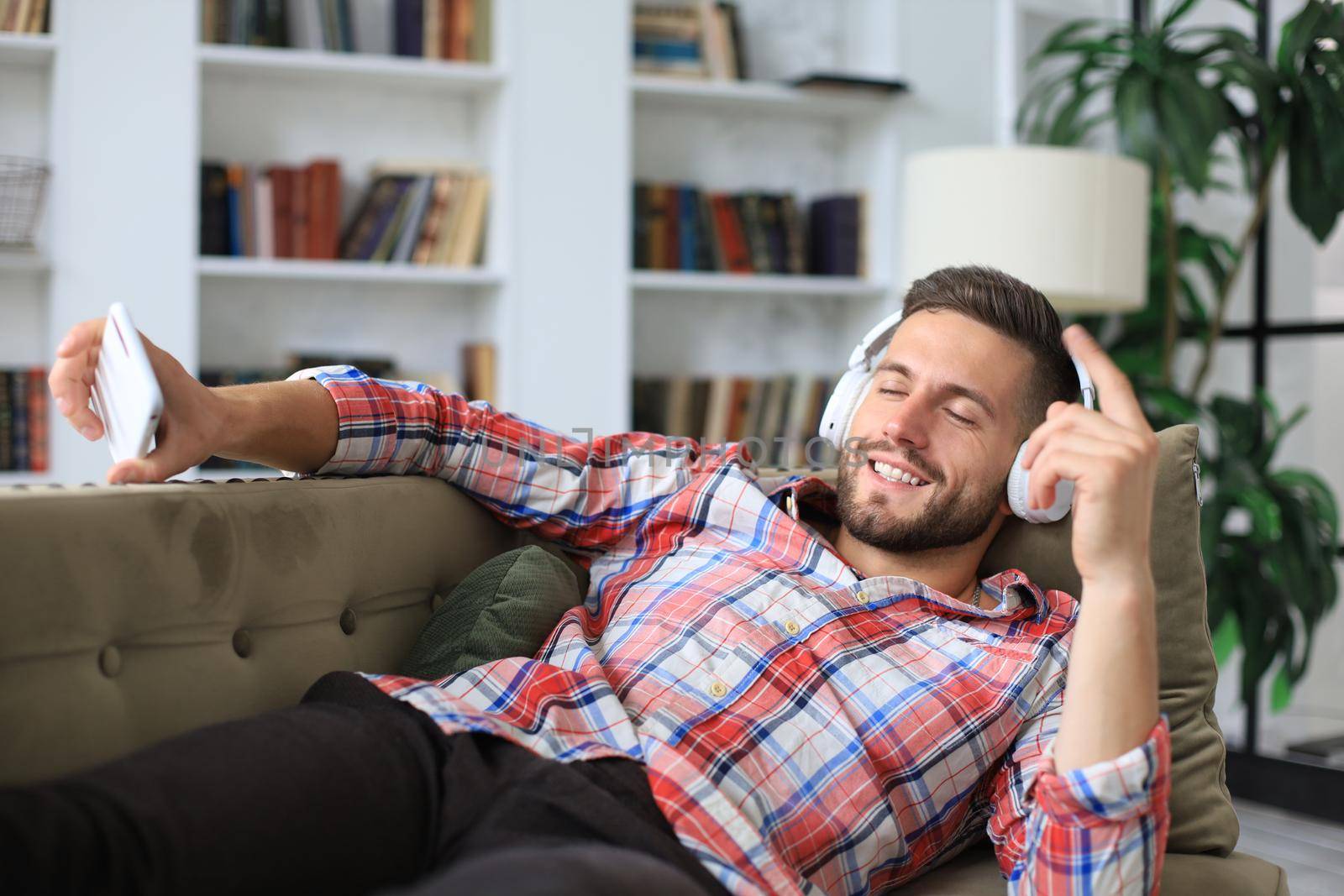 Attractive young man relaxing on a couch at home, listening music and playing on smartphone