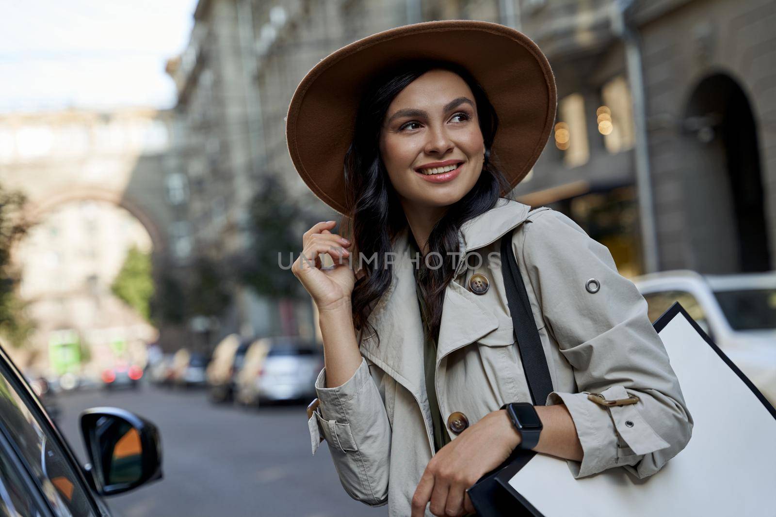 Portrait of a young attractive caucasian woman in hat with shopping bag looking aside and smiling while standing on the city street after visiting exclusive boutiques. Fashion, people lifestyle