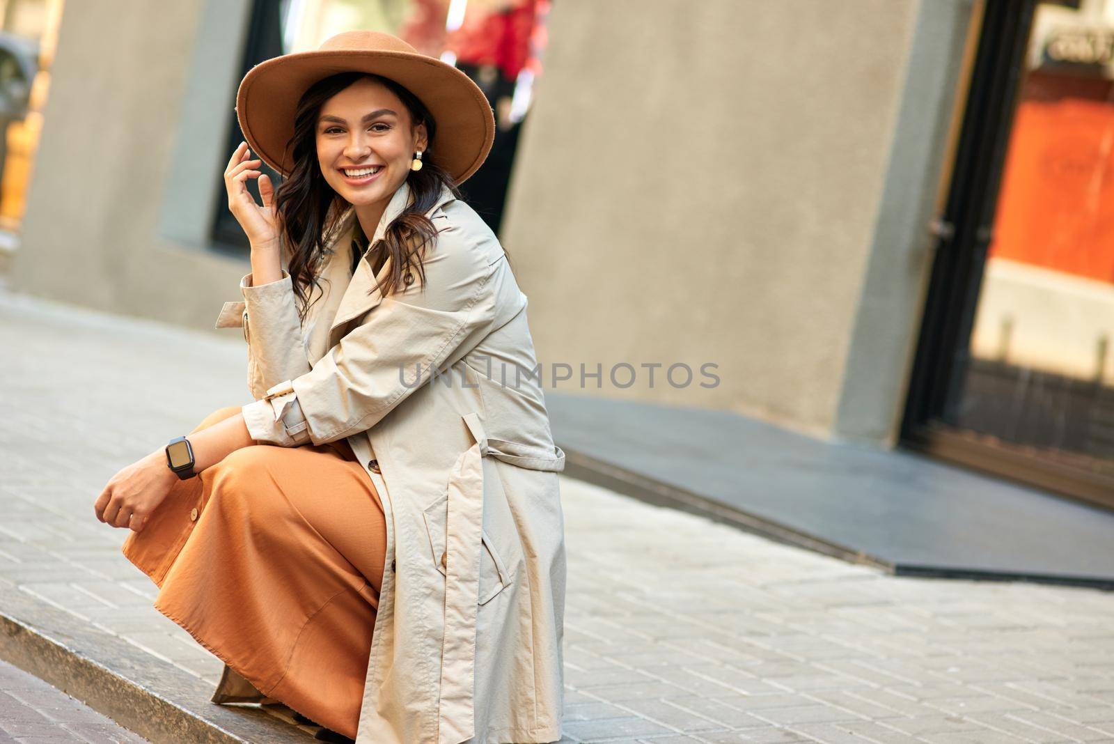 Enjoying nice warm day outdoors. Young attractive happy woman wearing autumn coat and hat looking at camera and smiling while walking city streets by friendsstock