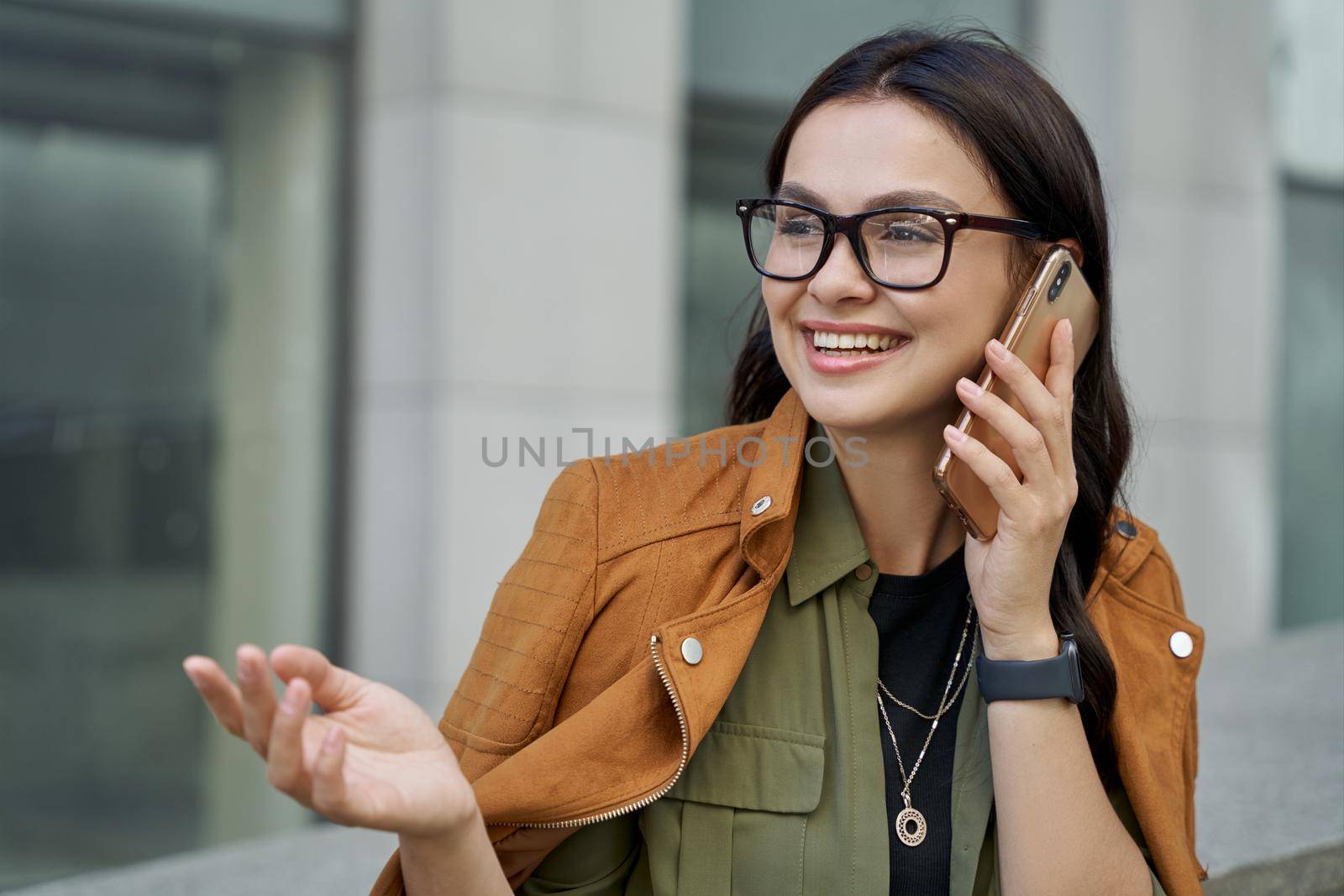 Always in touch. Portrait of a young beautiful happy business woman wearing eyeglasses talking on smartphone, looking aside and smiling while standing on the city street by friendsstock